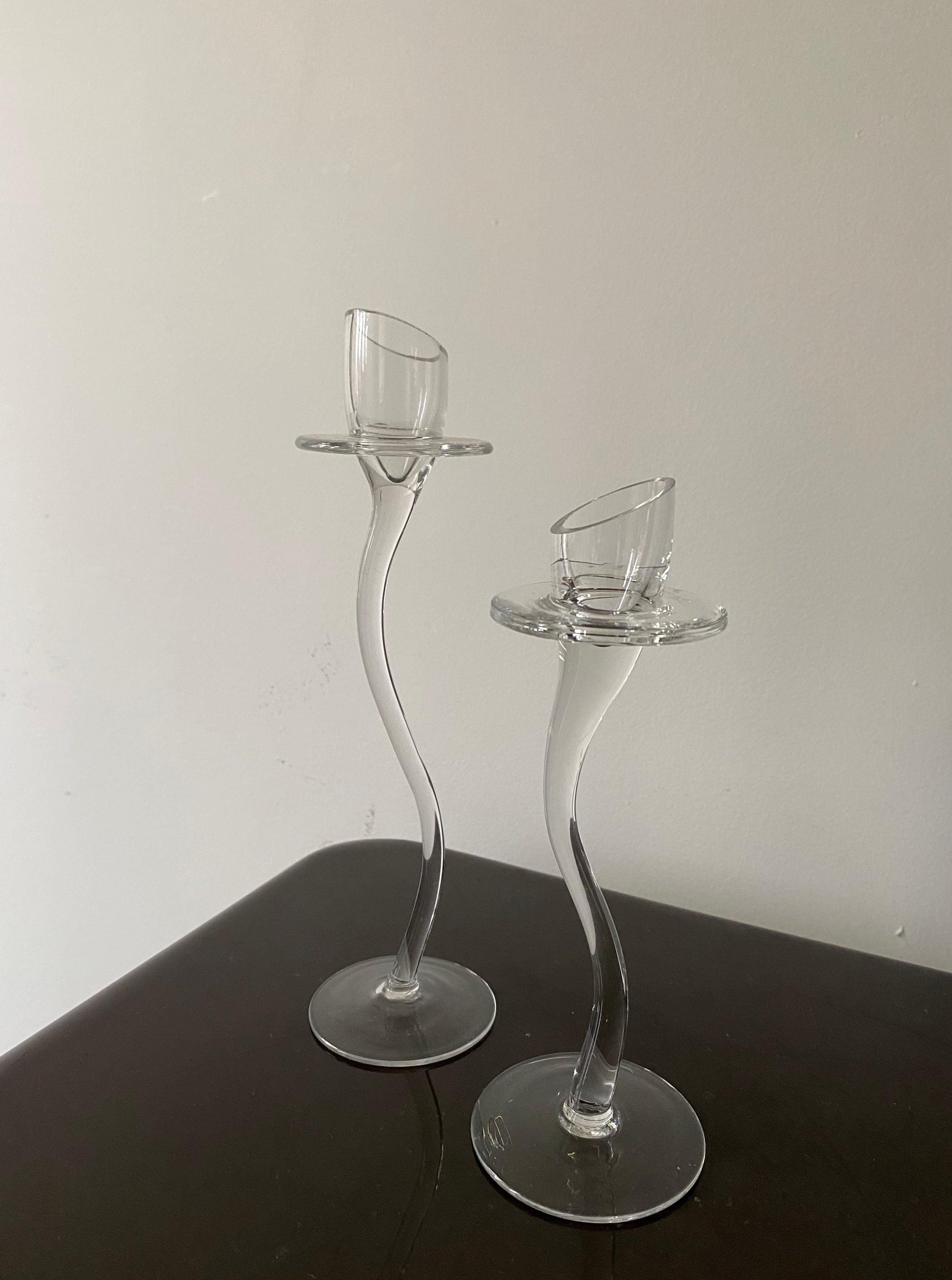 Two elegant, tall Les Objets Amsterdam vintage-inspired candle holders with twisted stems and glass holders, standing on a wooden surface against a plain light-colored wall.