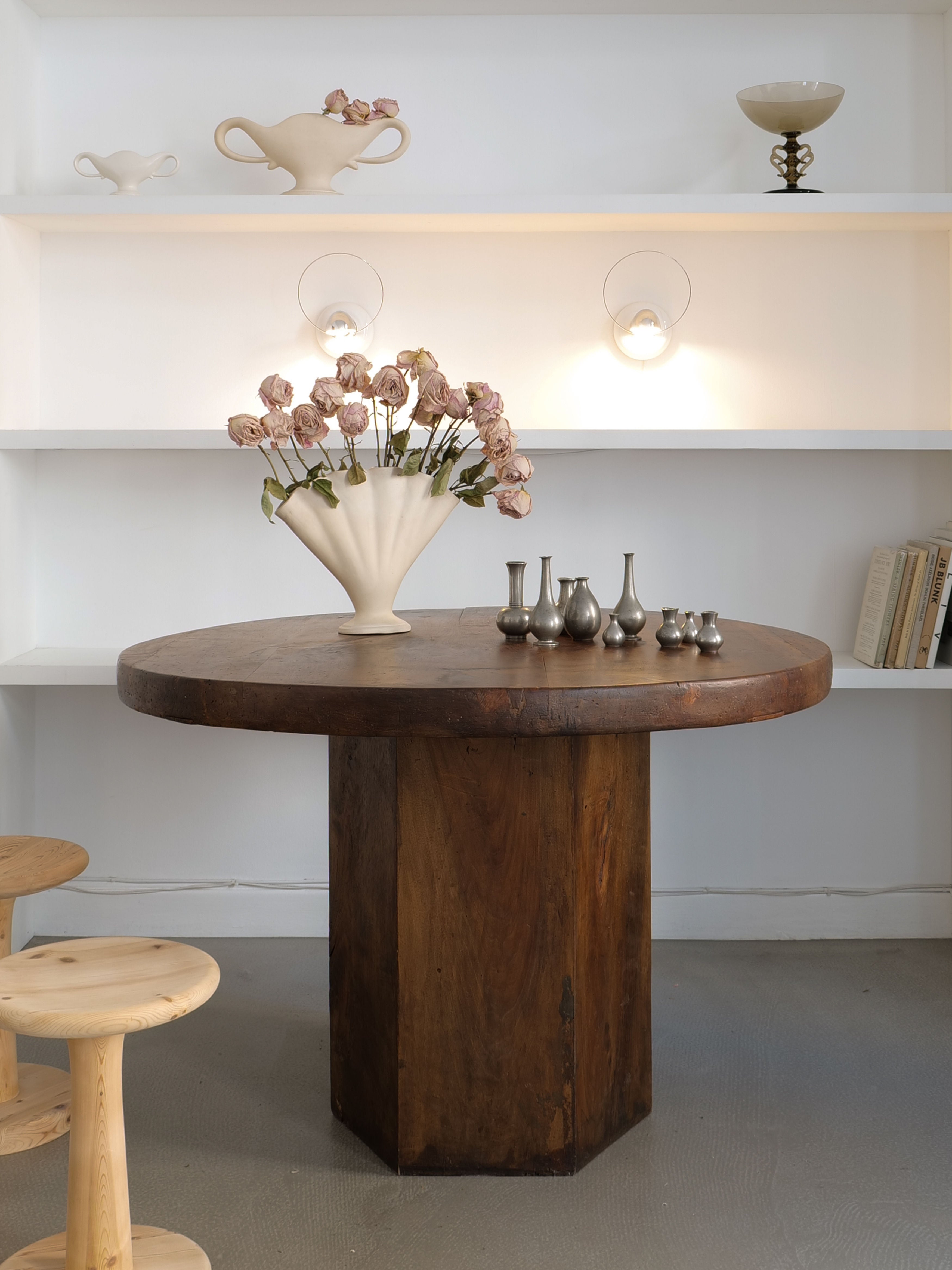 A wooden table with a fluted vase of dried flowers is centered beneath wall-mounted shelves displaying white ceramics. Small pewter vessels, including the exquisite pieces from the Just Andersen Mini Vase Collection by Collection apart, are lined up on the table. Two wooden stools are partially visible in the foreground, and a few books are on the shelf.