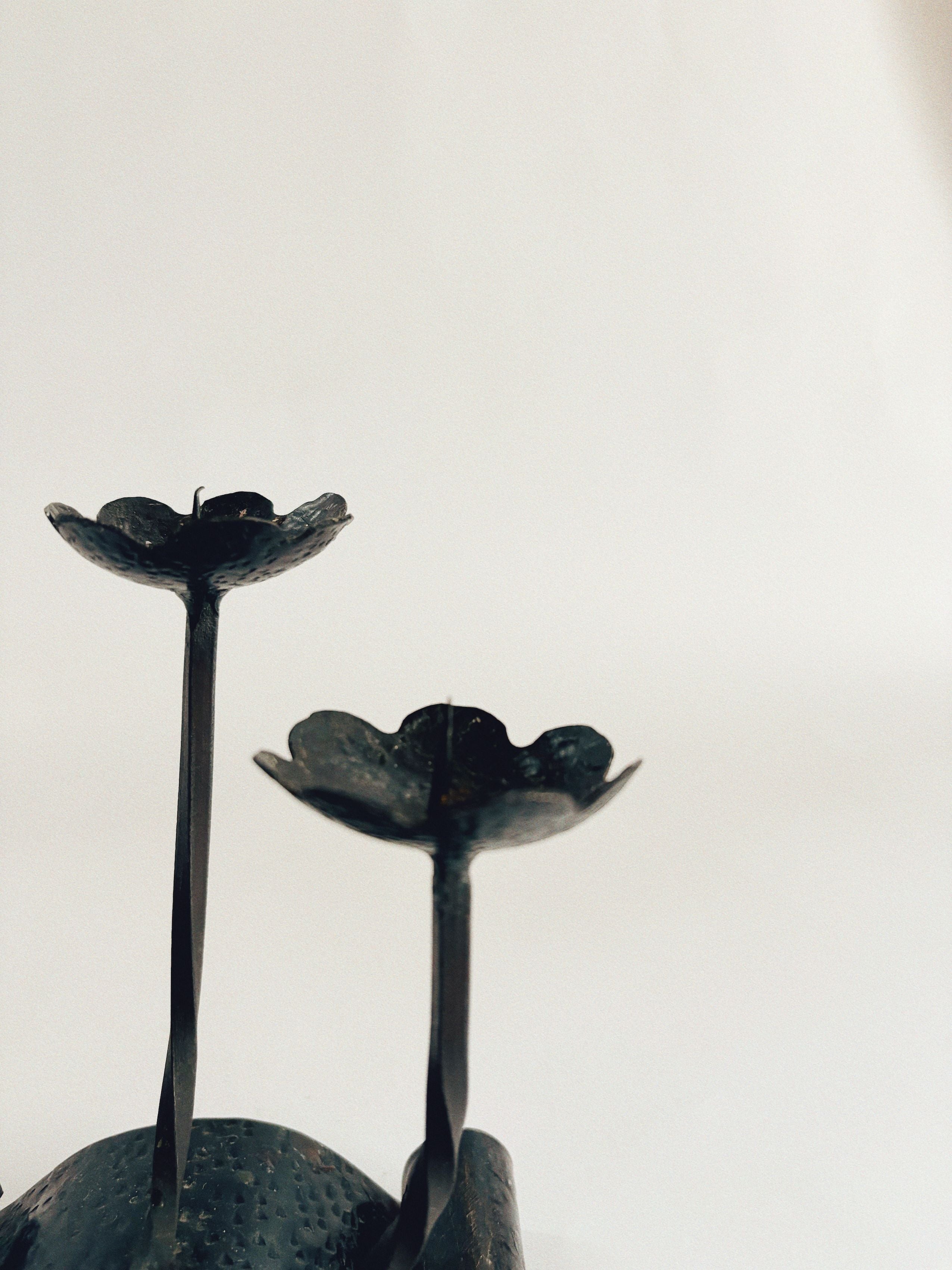 On the left side of the image, set against a simple, light backdrop, are two Handmade Metal Flower Candlesticks by Huerto featuring an artist-crafted design and a textured, vintage charm.