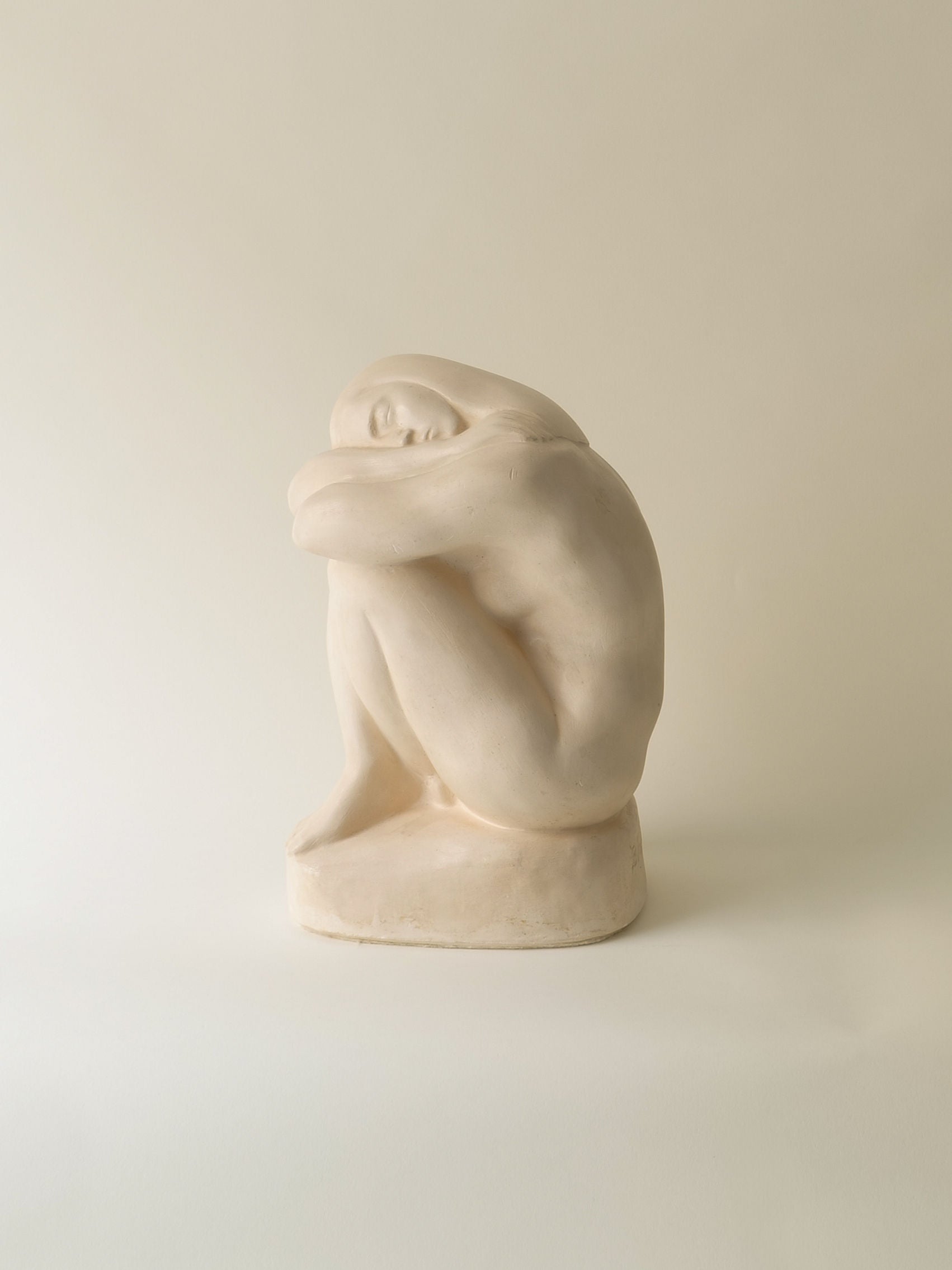 The "Resting Sculpture" by Collection Apart's Bror Forslund portrays a seated figure with their head on their knees and arms wrapped around their legs. Crafted in light hues, it epitomizes minimalistic elegance against a simple, neutral backdrop.