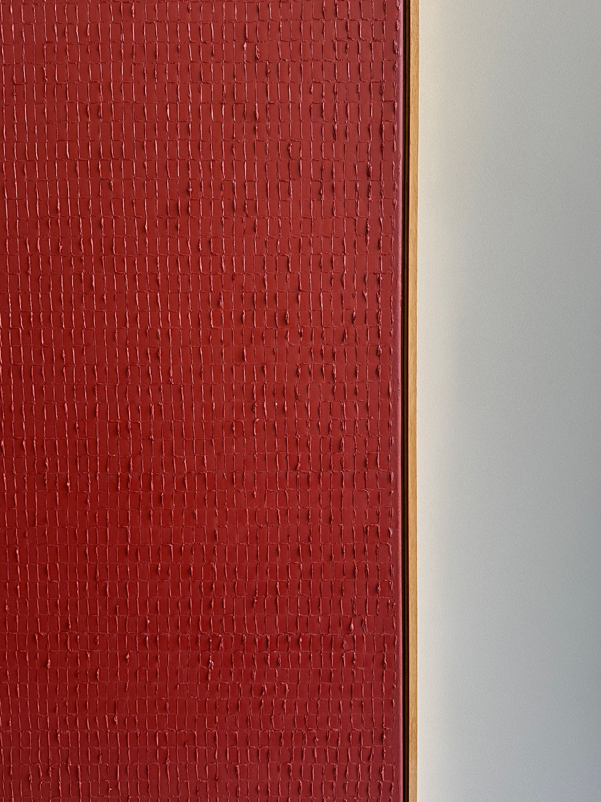 A close-up of a wall featuring a red textured surface on the left and a smooth white surface on the right. The textures, reminiscent of the Dansaekhwa movement, create a striking contrast between the two sections, separated by a narrow wooden trim. This visual interplay invites mindfulness in its observation and is evocative of *Indian Red - Oil on canvas* by *Roi Elmaliah*.