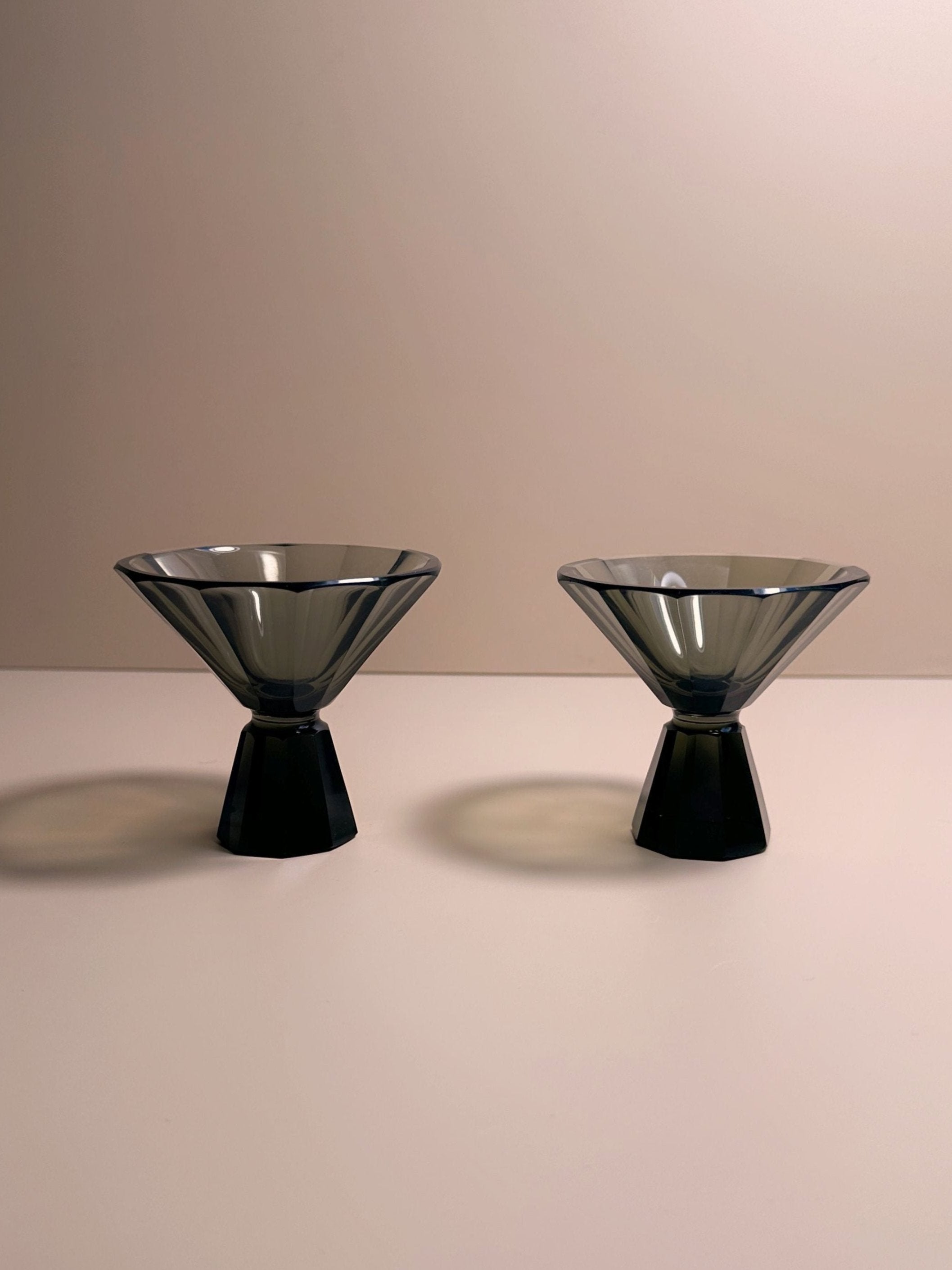 Two sleek, modern, 1930s Art Deco Facetted Liqueur Glasses by Dodo Vintage with wide rims and narrow bases are symmetrically positioned side by side on a smooth, light-colored surface against a simple, beige background. These handmade pieces cast soft shadows beneath them.