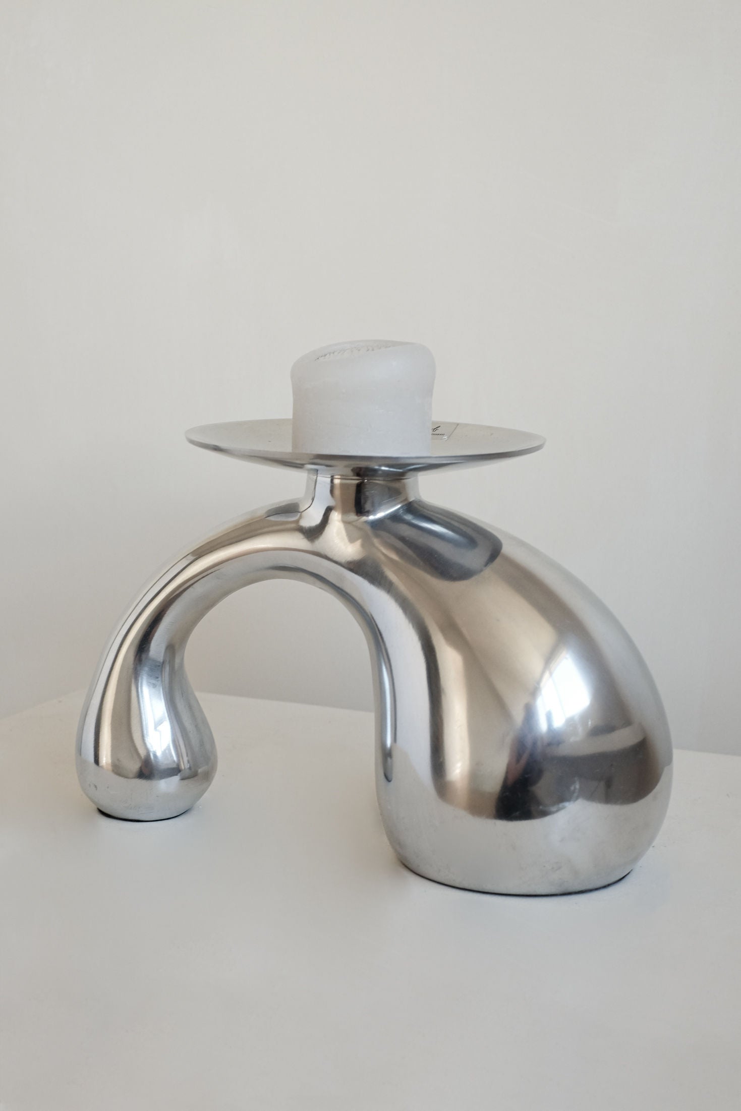 The Bombastic ‘Drop’ Candle Holder by septembre studios features a sculptural, polished metal arched base with a frosted white candle, perfect for modern interiors and shown against a white background.