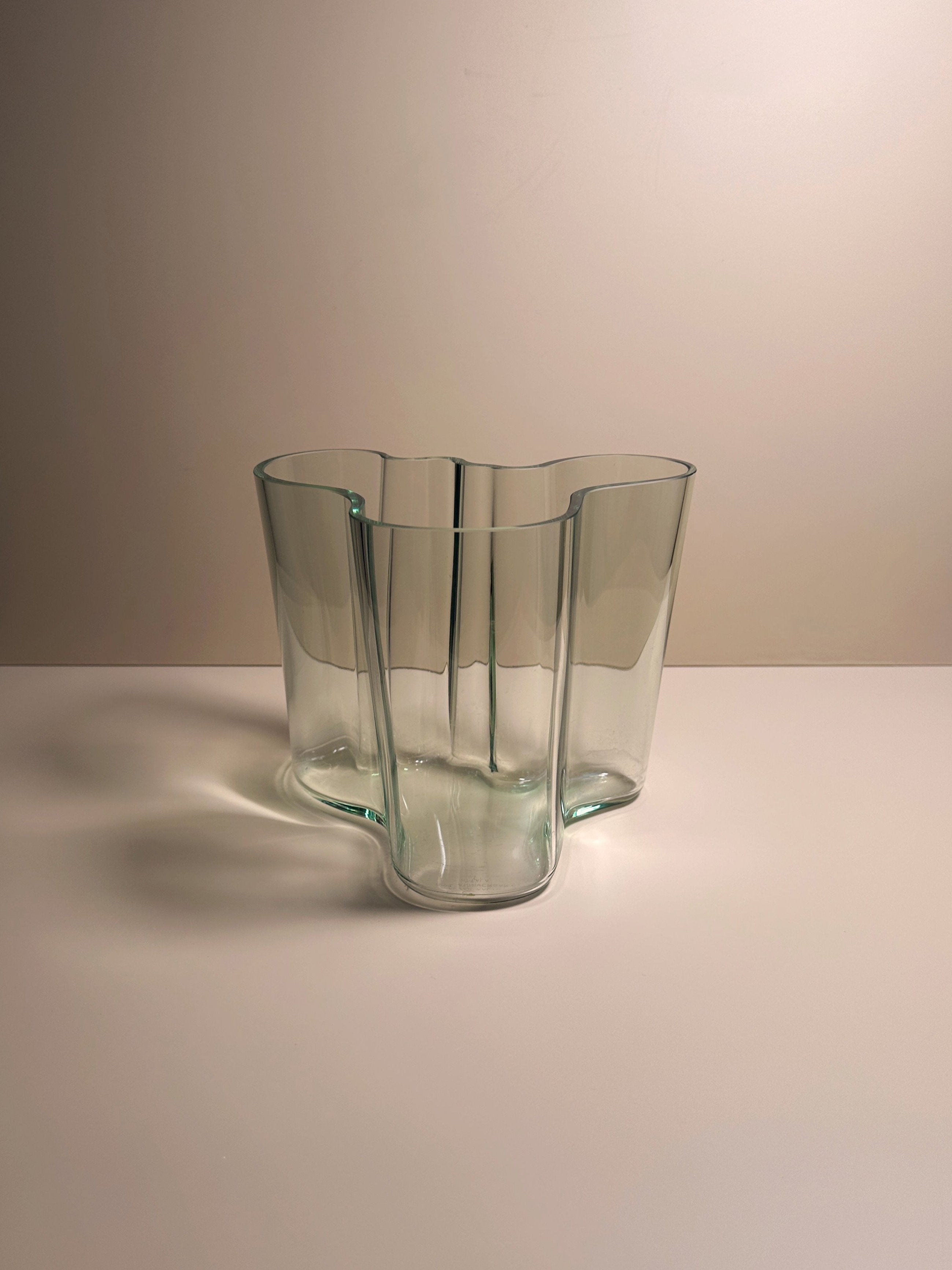 A Limited Edition Aalto Vase by Dodo Vintage, featuring a transparent, wavy-edged and uniquely undulating design, is centered on a plain, light-colored surface against a neutral wall background. This collectible piece casts soft shadows on the surface below it.