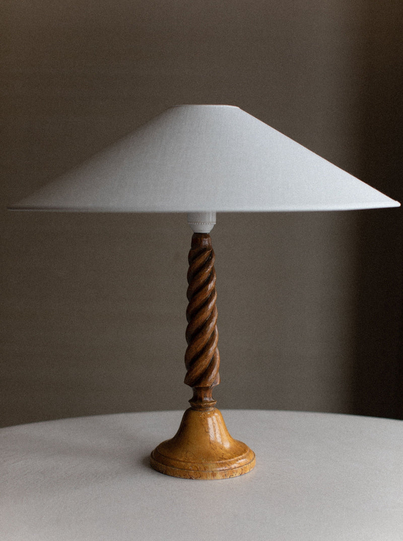 A Twisted Wooden Lamp by Out For Lunch with a wide, white conical lampshade and a twisted base stands on a round white table. The natural wood grain showcases intricate spiral carvings, highlighting its artisanal craftsmanship. The background is a neutral, blurred wall.