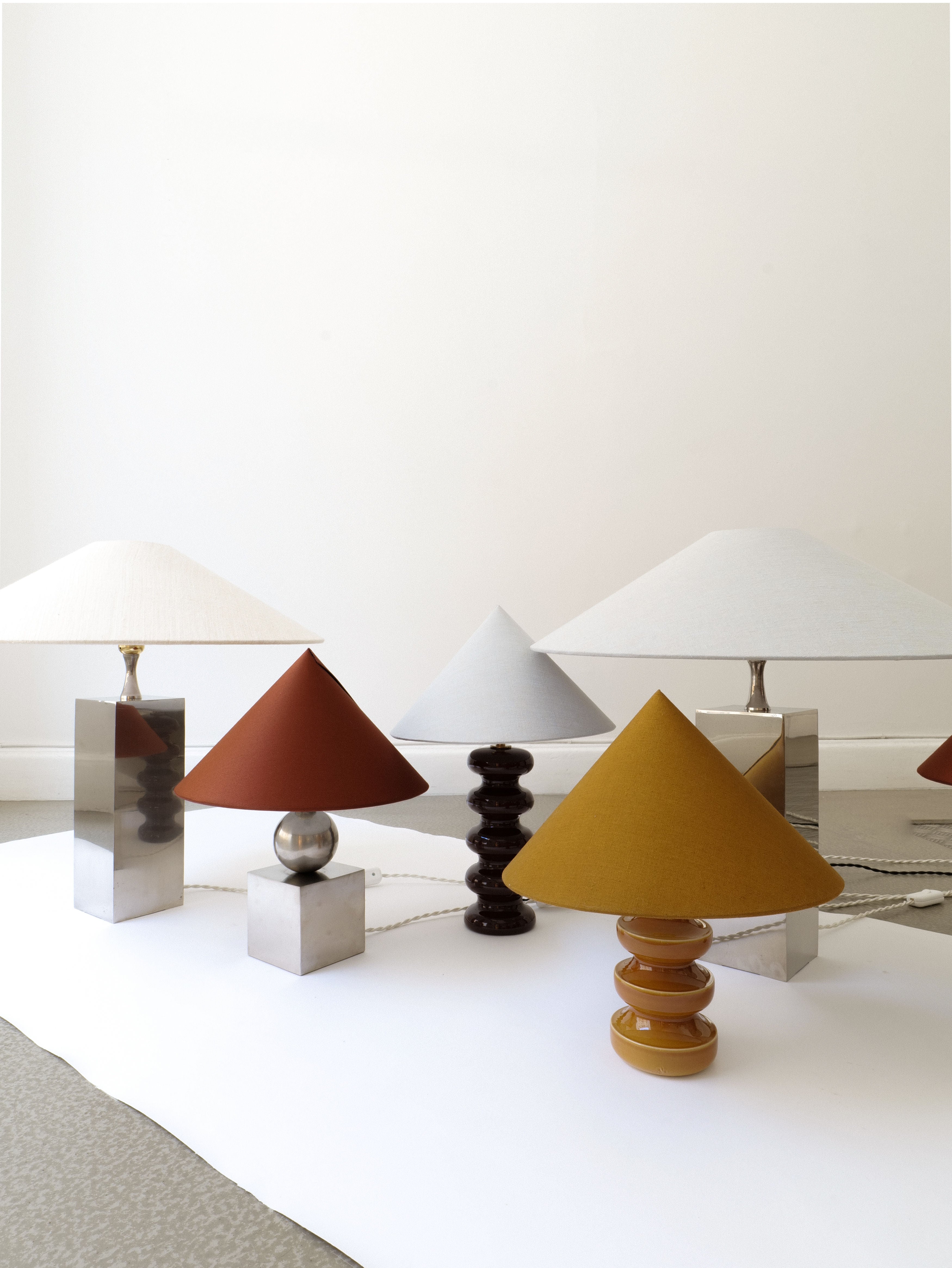 A collection of modern table lamps, some with brass fittings, is displayed against a plain white wall. Featuring various geometric shapes and colors, the display includes silver bases with white and orange shades alongside a mustard yellow Small Sculptural Lamp by Collection apart with a cylindrical stacked base.