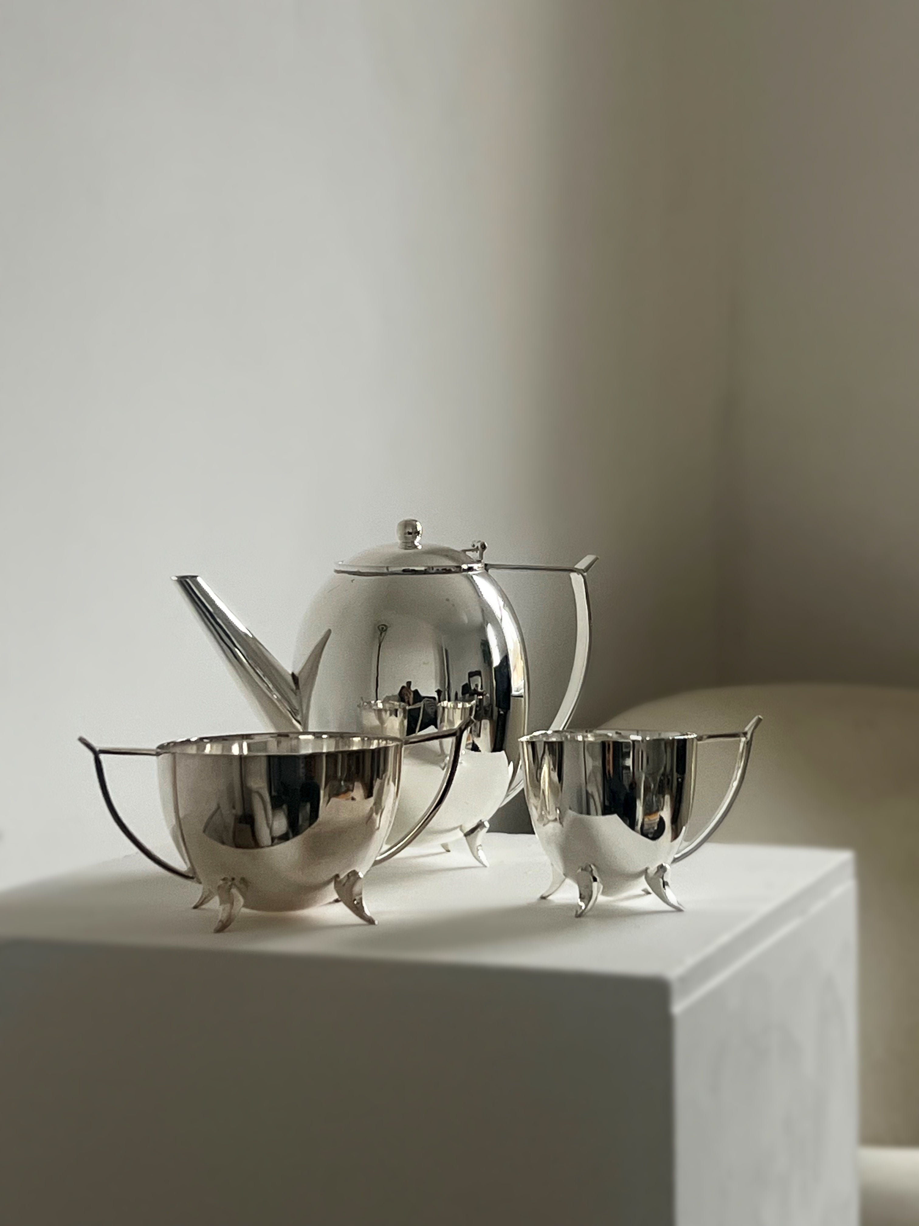 The Porter Studio "Elegant Three Piece Tea Set," a polished silver-plated set with deco feet, including a teapot and two sugar bowls, sits elegantly on a white pedestal against a soft gray background, glistening as it reflects the light to highlight its smooth, shiny surface.