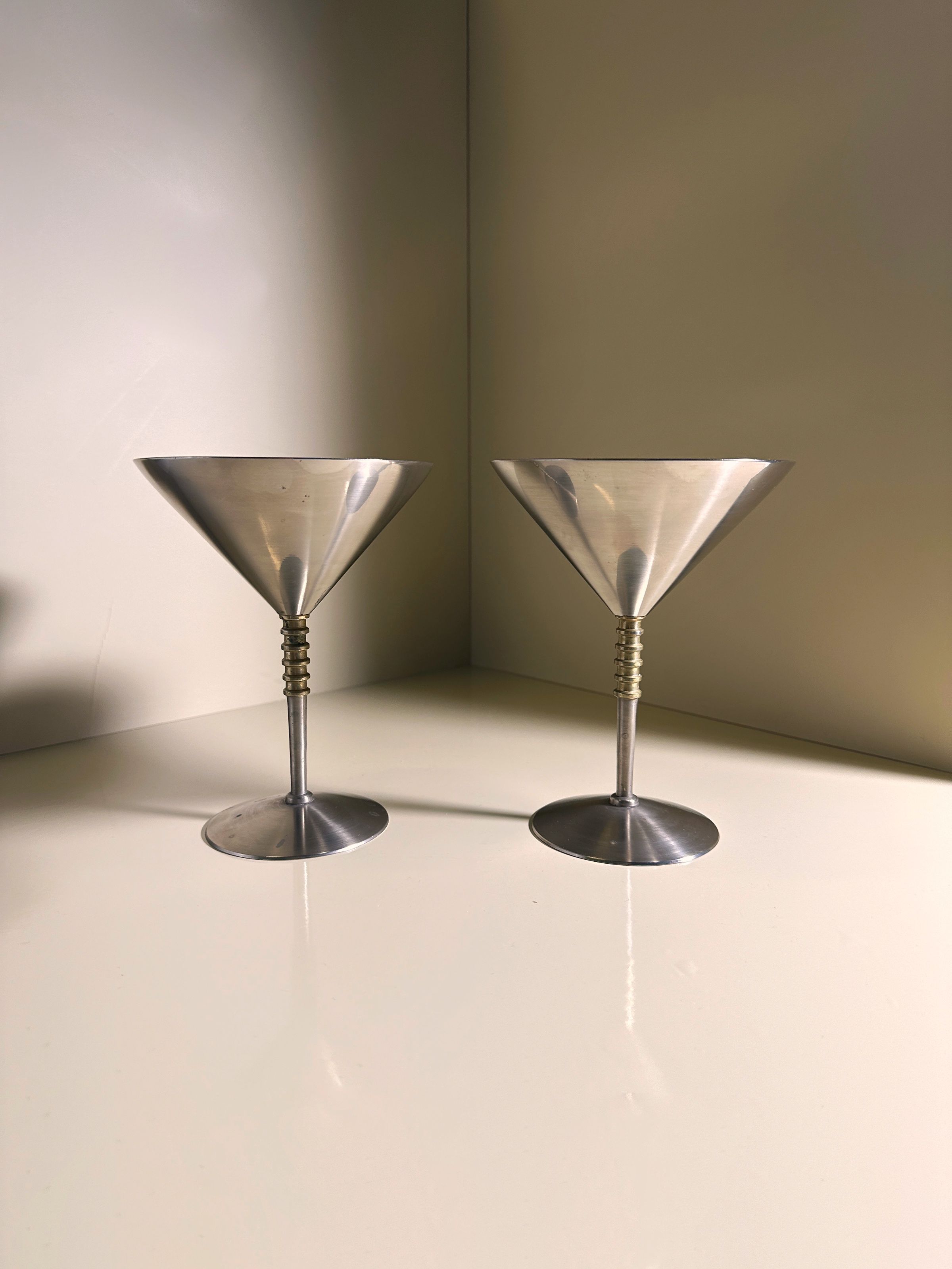 Two solid metal coupés with brass details from Dodo Vintage rest on a glossy surface, against smooth, light-colored walls that cast soft reflections around the glasses.