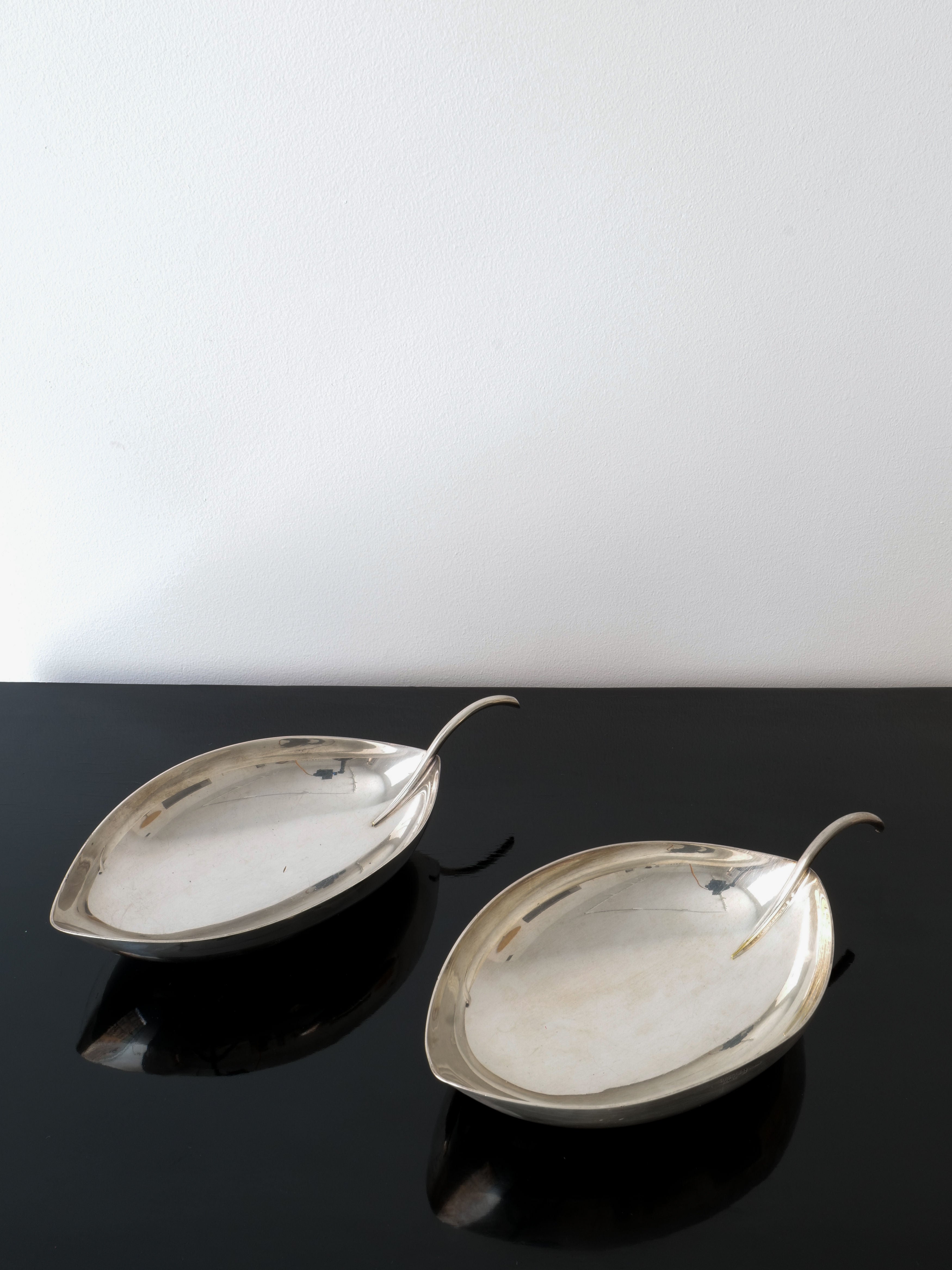 Two Christofle Leaf Vide Poche Lino Sabattini serving dishes from Collection Apart, with their elegant leaf-shaped design and distinctive curved handles, sit gracefully on a glossy black surface against a white background. Their simple and sophisticated form casts subtle reflections on the surface below.