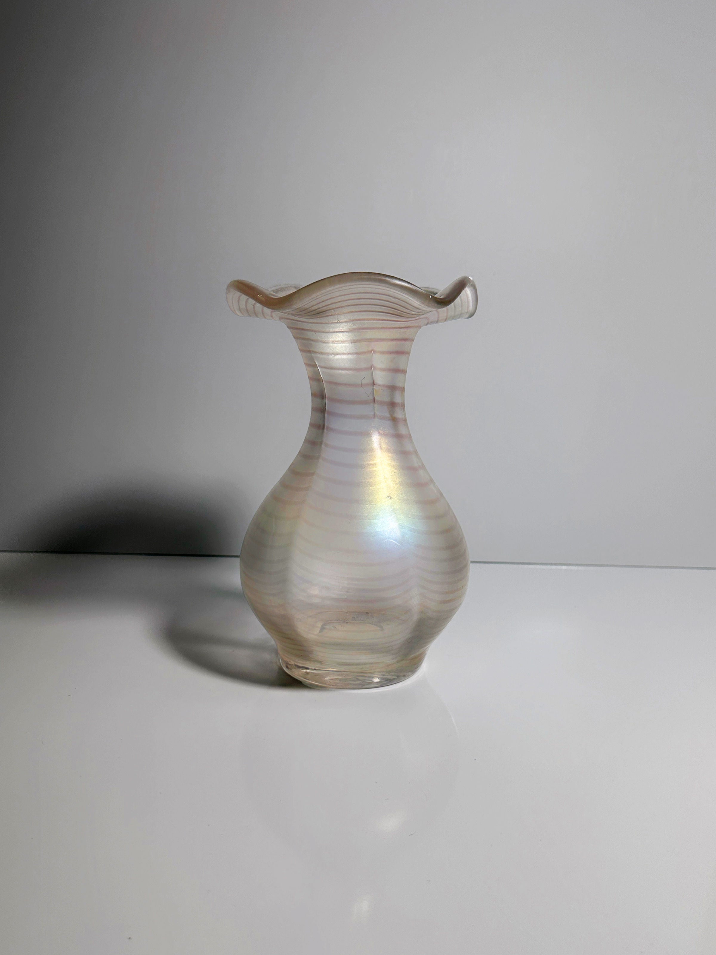 The Art Nouveau Wavy Vase by Dodo Vintage is a translucent, pear-shaped iridescent glass vase that beautifully highlights soft rainbow colors. Its elegant flared, wavy rim rests gracefully on a smooth white surface set against a pale gray background, showcasing the timeless charm typical of Gallo design.