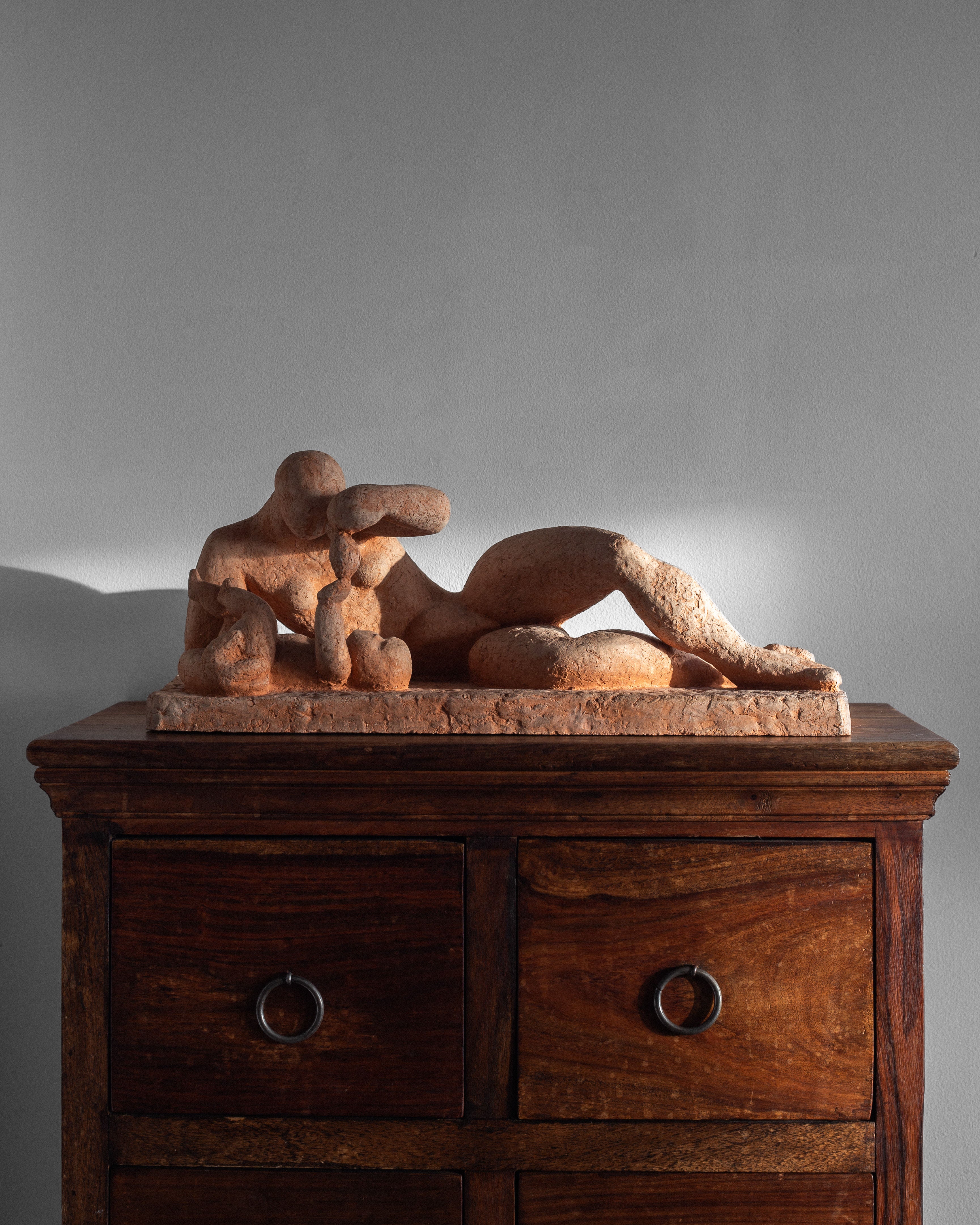 On a two-drawer wooden cabinet sits a Modernist Terracotta Sculpture by Bengt Amundin from the 1950s, offered by Spigel. Soft lighting casts gentle shadows on the wall behind, evoking Swedish modernist art's elegance.