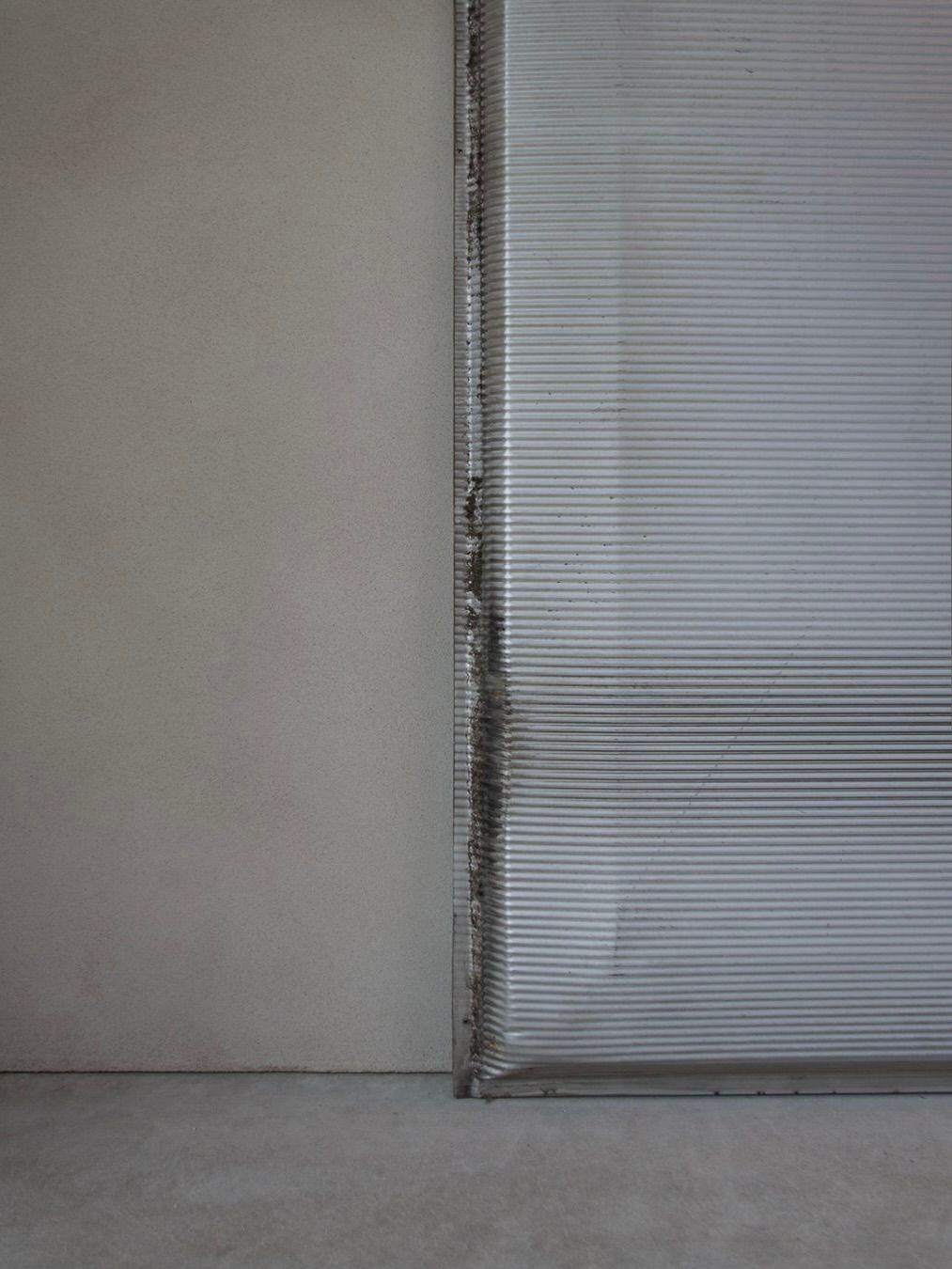 A partially closed, dented metal shutter resembling the Jean Prouvé - Façade Panel 108 by Bicci de' Medici contrasts against a plain beige wall. The concrete floor and shadows highlight the interplay between the wall and shutter, emphasizing its wear and textured panels.