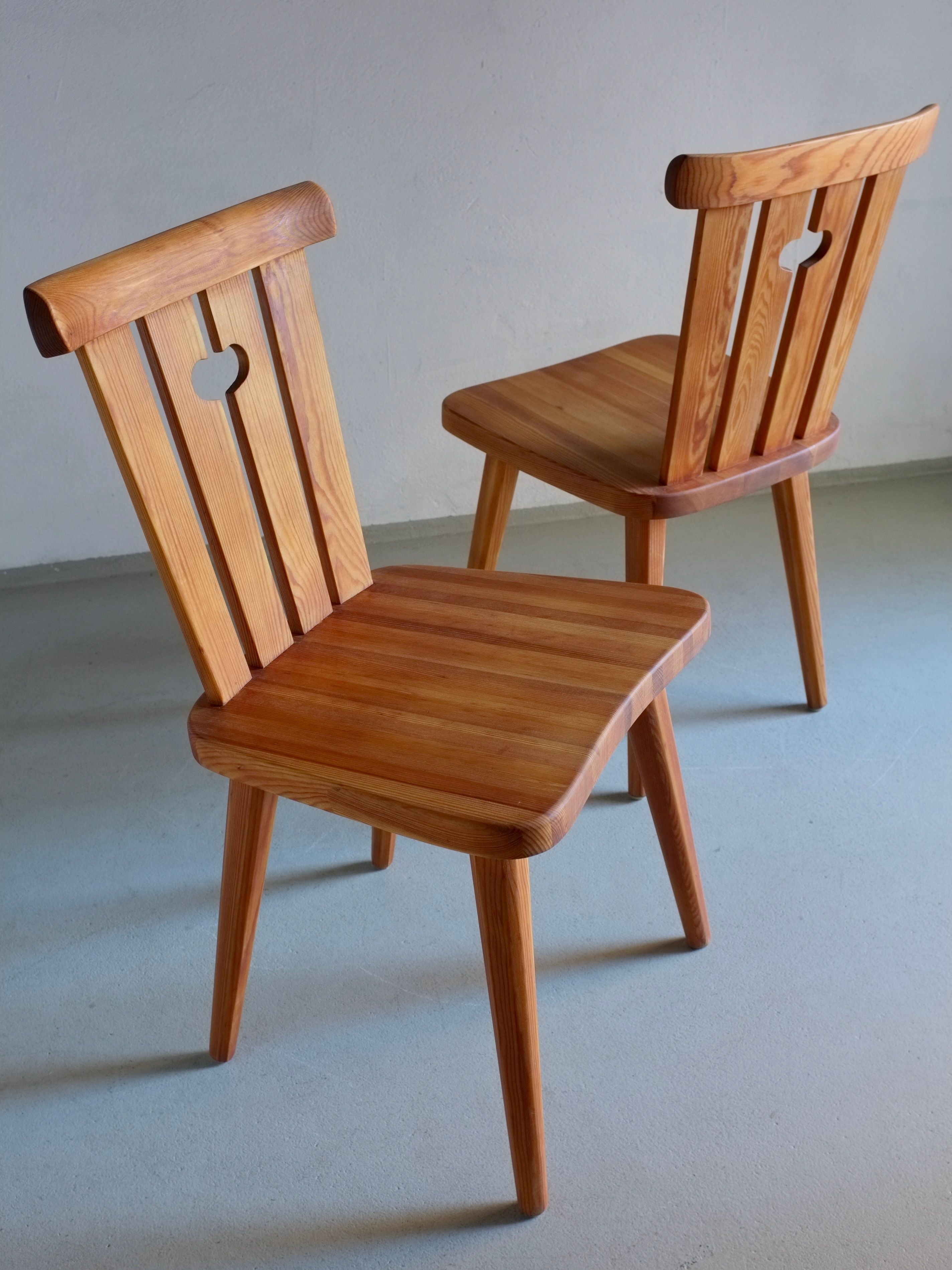 A pair of restored 1940s rustic pine chairs, created by Göran Malmvall for Veter Vintage, are set against a light gray floor. These chairs showcase slatted backs, rounded seats, and a heart-shaped cutout in the backrest. Smoothly finished with oil treatment, they are positioned slightly facing away from each other.