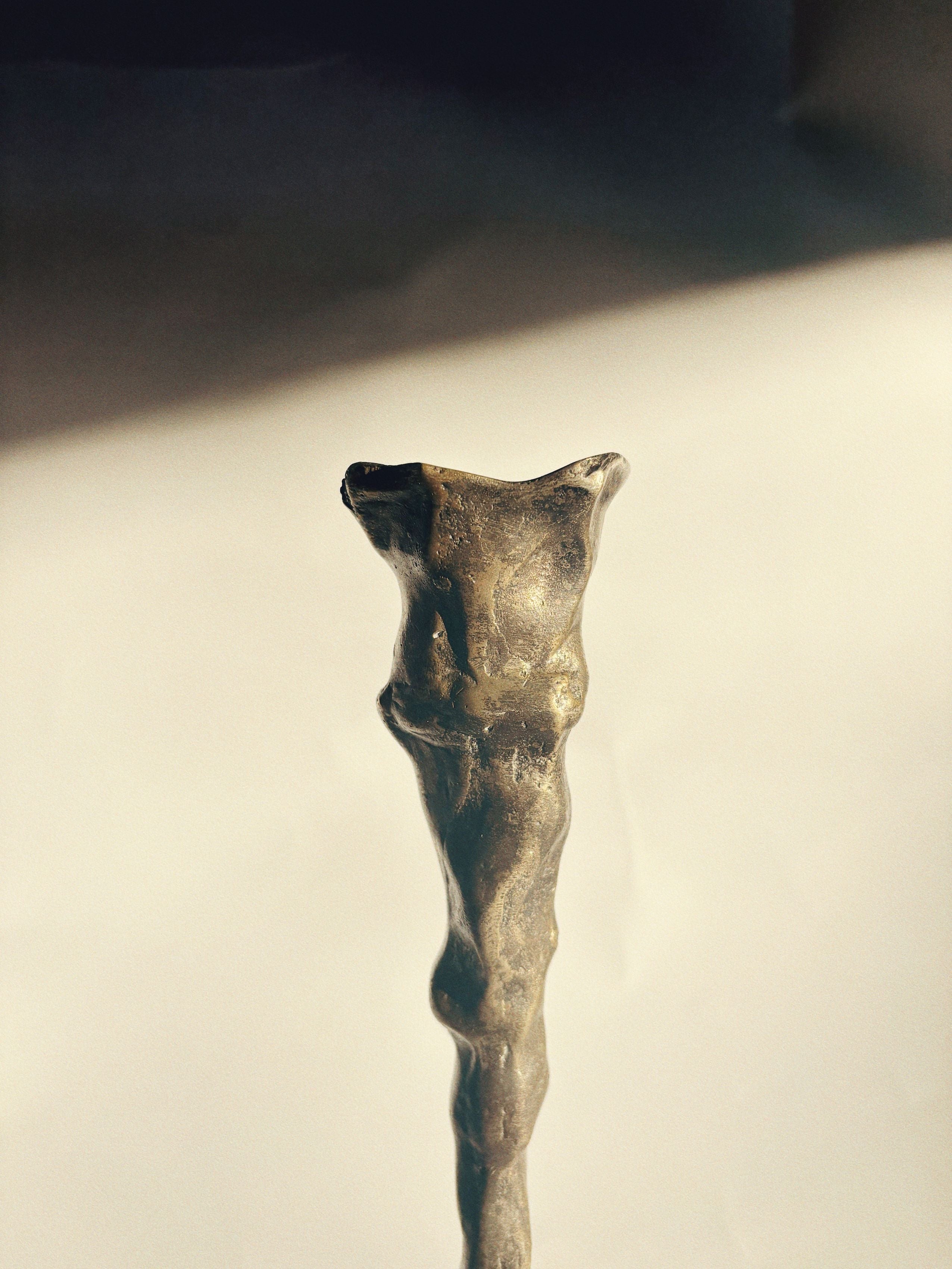 Close-up of the Huerto Bronze Candle Holder showcasing its abstract, elongated human-like form against a softly lit background. This distinctive piece features a rough, organic texture and is artist-signed, casting subtle shadows reminiscent of fine artistry.