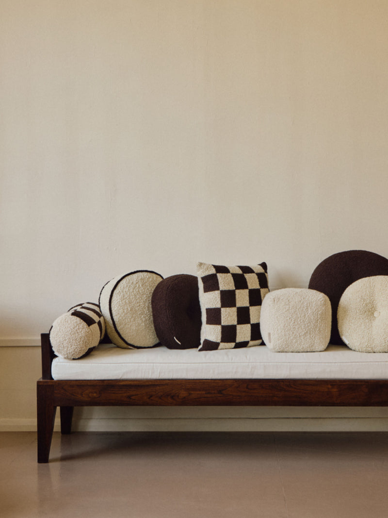 A minimalist seating area featuring a wooden-framed bench with a white handmade cushion. The bench is adorned with various plush pillows in black and white, including checkered, round, and cylindrical shapes made of British eco wool, including AMÉLIORÉ’s Le Carré - Wool Bouclé - Chocolate/Eggshell, creating a cozy and stylish arrangement against a plain beige wall.