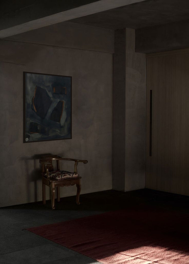 A dimly lit room features a wooden chair against a wall. Above the chair hangs "Complex Hue's in G Minor," a dark, abstract mixed media piece on linen by Yeliza Gevorgyan, in an Italian oak frame. The floor is partially covered by a dark rug, and to the right, a large wooden door casts shadows that create a serene and moody atmosphere.