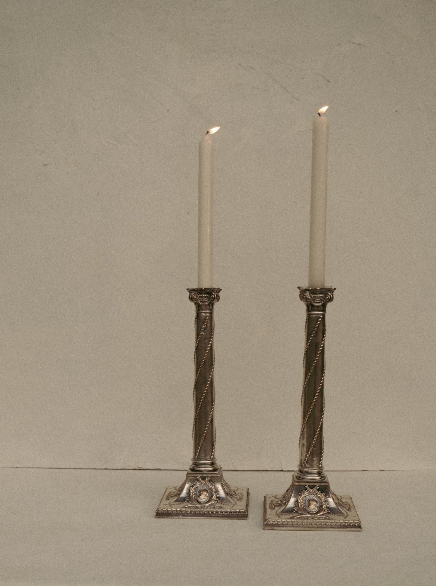 Two ornate Casita Amada Pair of Column Sheffield silver plated candle stick holders with intricate detailing, each holding a lit white taper candle, stand side by side in antique condition on a plain surface against a beige background.