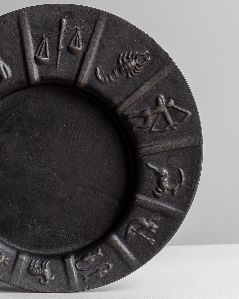 The Spigel Cast Iron Zodiac Dish showcases a vintage appeal with symbols of the zodiac, including Libra, Scorpio, and Sagittarius, gracefully decorating its black ceramic rim. Set against a neutral backdrop, the dish emanates timeless charm that echoes the craftsmanship of Ystad Metall.