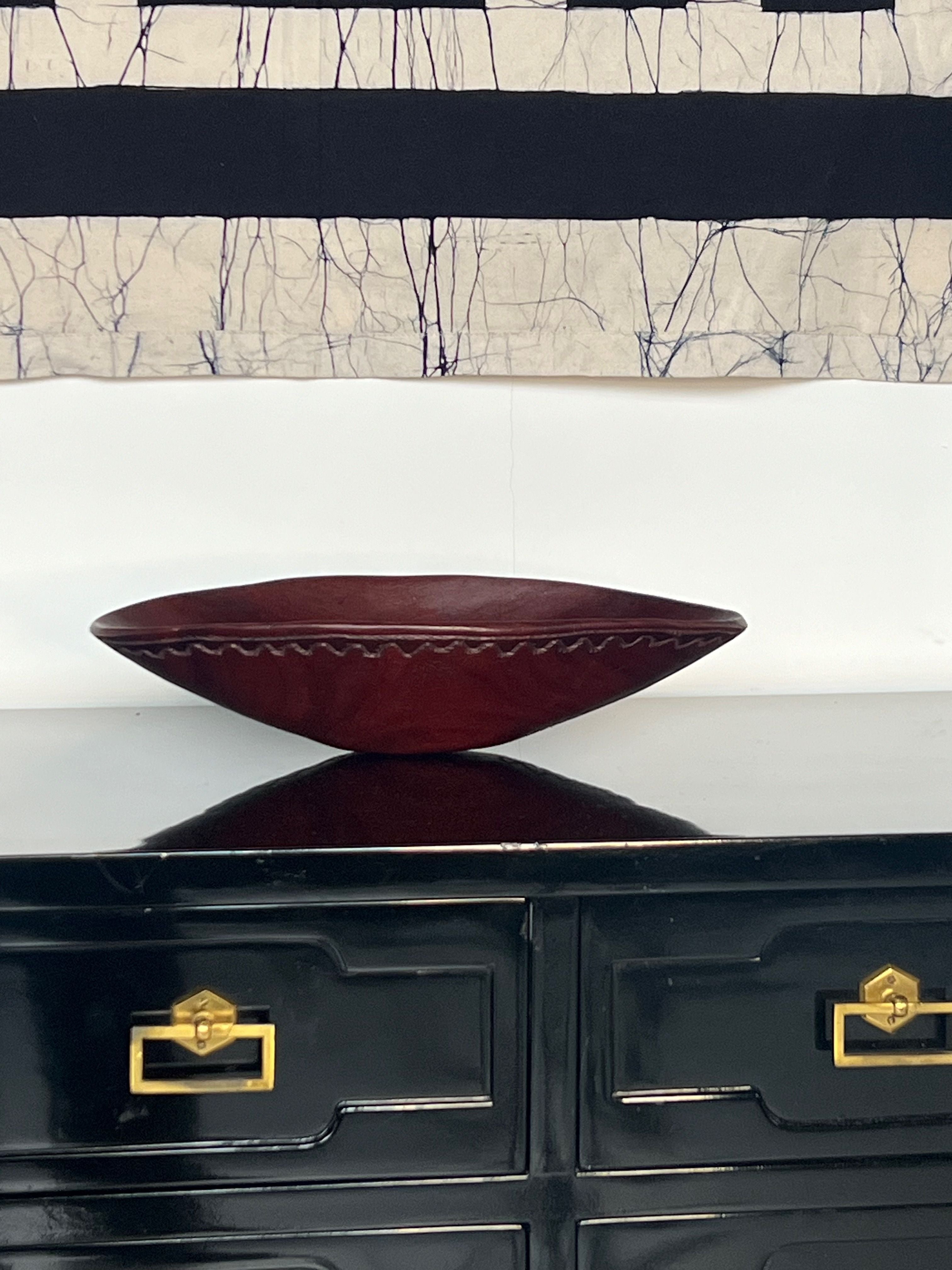 A sleek Carved Wooden Bowl from Porter Studio sits on a glossy black dresser with gold handles. Above, a decorative textile featuring abstract geometric patterns in black and white hangs on the wall.