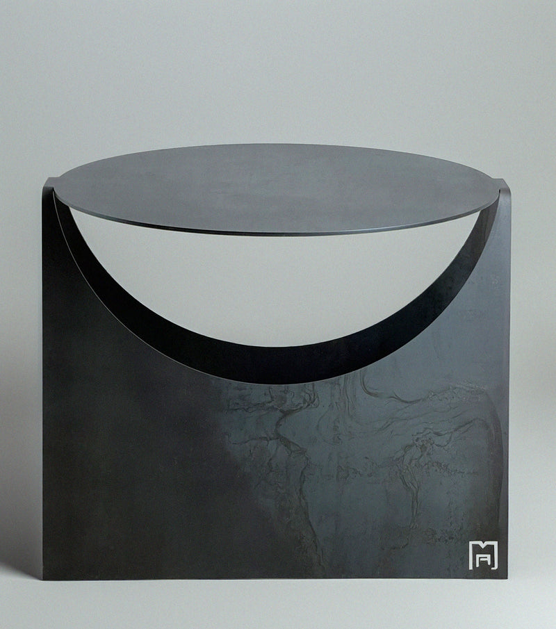 The 'Maru' Side Table by Modan is a minimalist black steel table with a round top, semicircular cutout beneath, and a small geometric logo in the lower right corner, set against a plain light gray background.