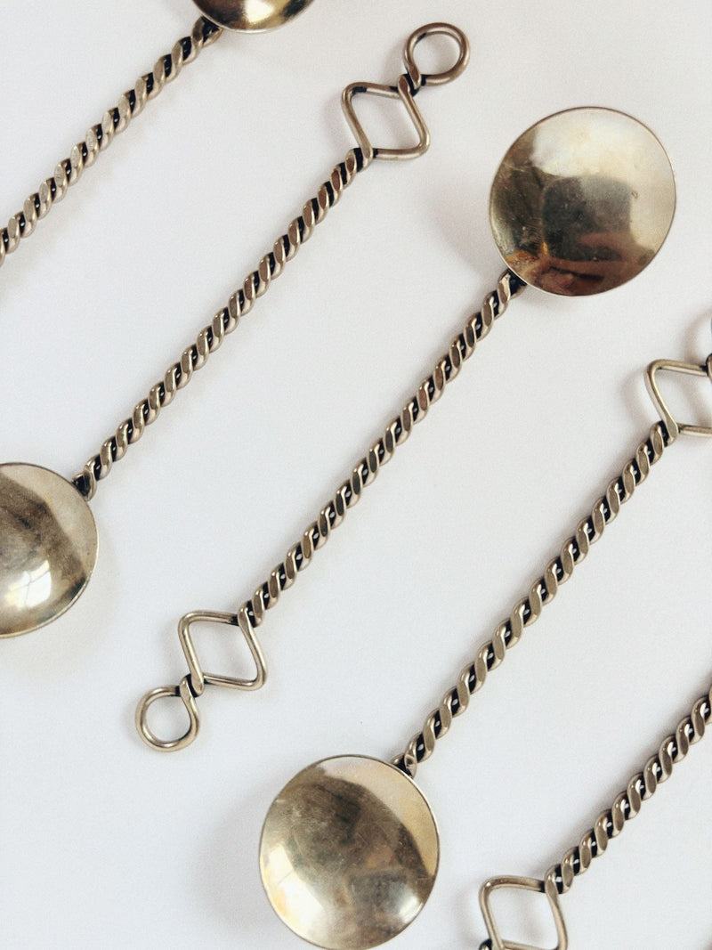 Close-up of Huerto's 70's Handmade Spoons Set with twisted handles and looped ends, displayed diagonally on white. These spoons showcase a reflective metallic surface and unique geometric designs, capturing the essence of functional art.