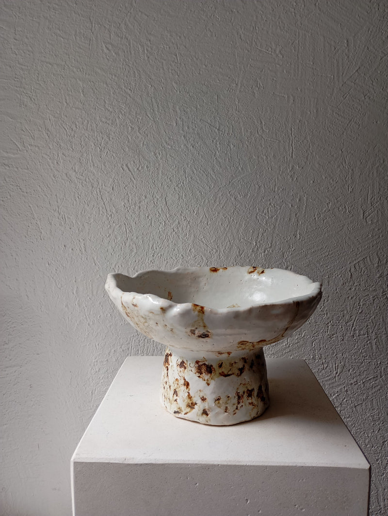 The Coupe 53 by Claire Pain is a stoneware bowl with a rough, irregular surface and white glaze with brown accents, displayed on a white pedestal against a textured gray wall.