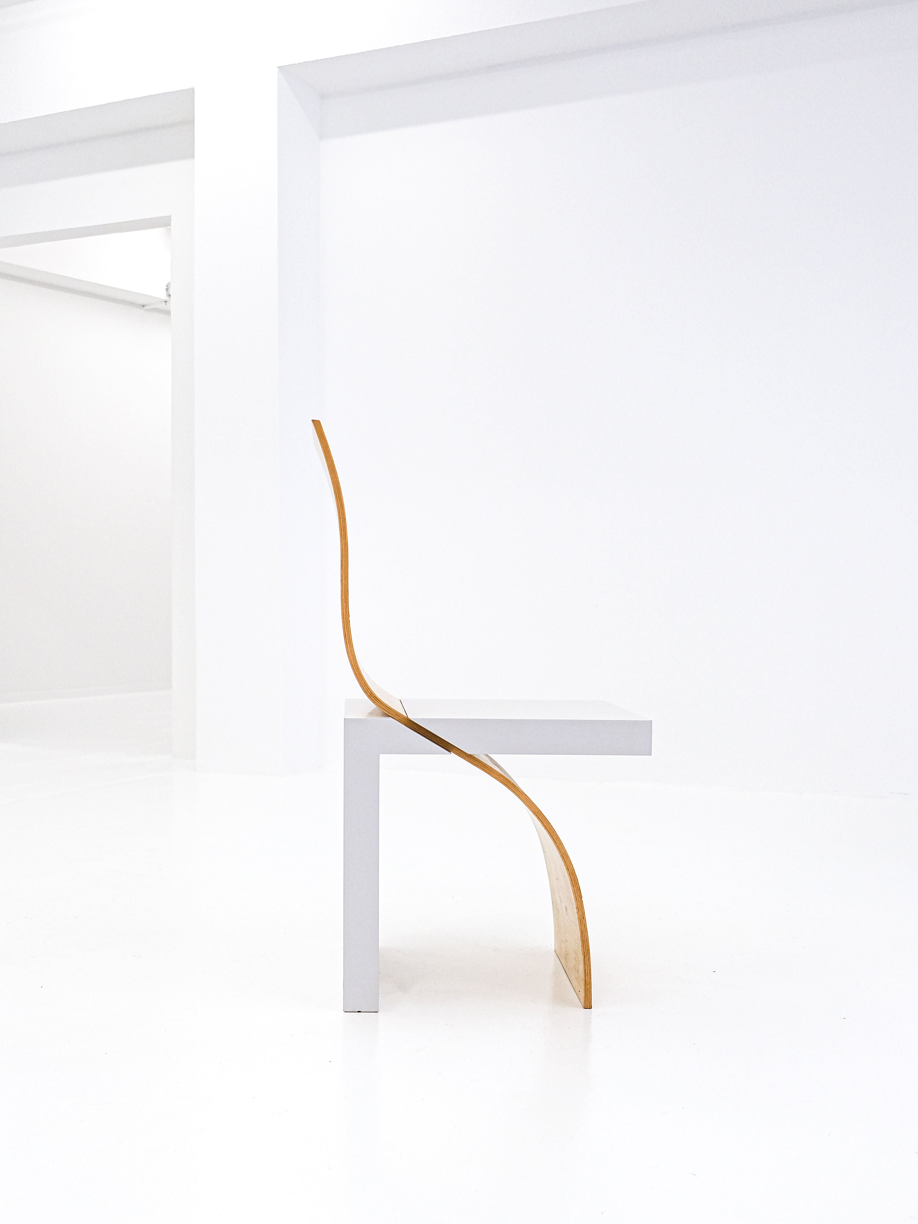 The Postmodern Leda Chair by Angela Oedekoven, produced by antibeige in 1990, features a minimalist design with its flat white seat and back supported by a thin curved wooden frame. It complements the stark white room's clean lines and spacious aesthetic.