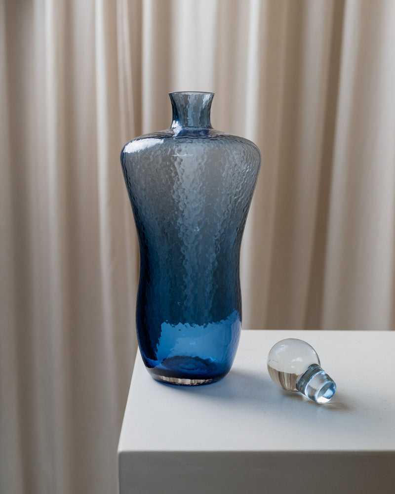 A Bottega Jacobs Vintage Glass Decanter in Blue, with a narrow neck and textured design, sits on a white pedestal alongside its detached stopper. The Hollywood Regency-inspired backdrop displays soft beige curtains.