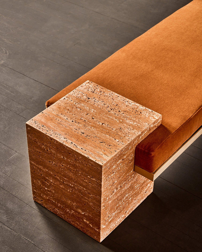 A close-up of the 'Coexist' Bench in Orange Travertine by Slash Objects features a rust-hued cushioned seat and a square orange travertine end, placed on a dark wood floor.