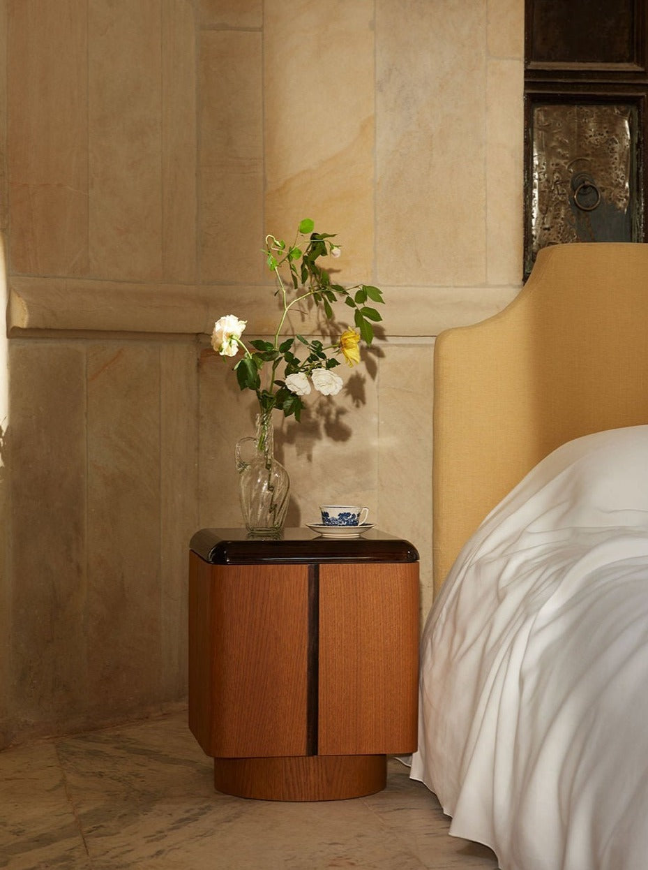 A cozy bedroom corner featuring a circular polished walnut Lemon nightstand with a clear vase holding delicate white and yellow roses, beside a plush bed with a light beige headboard and crisp white sheets.