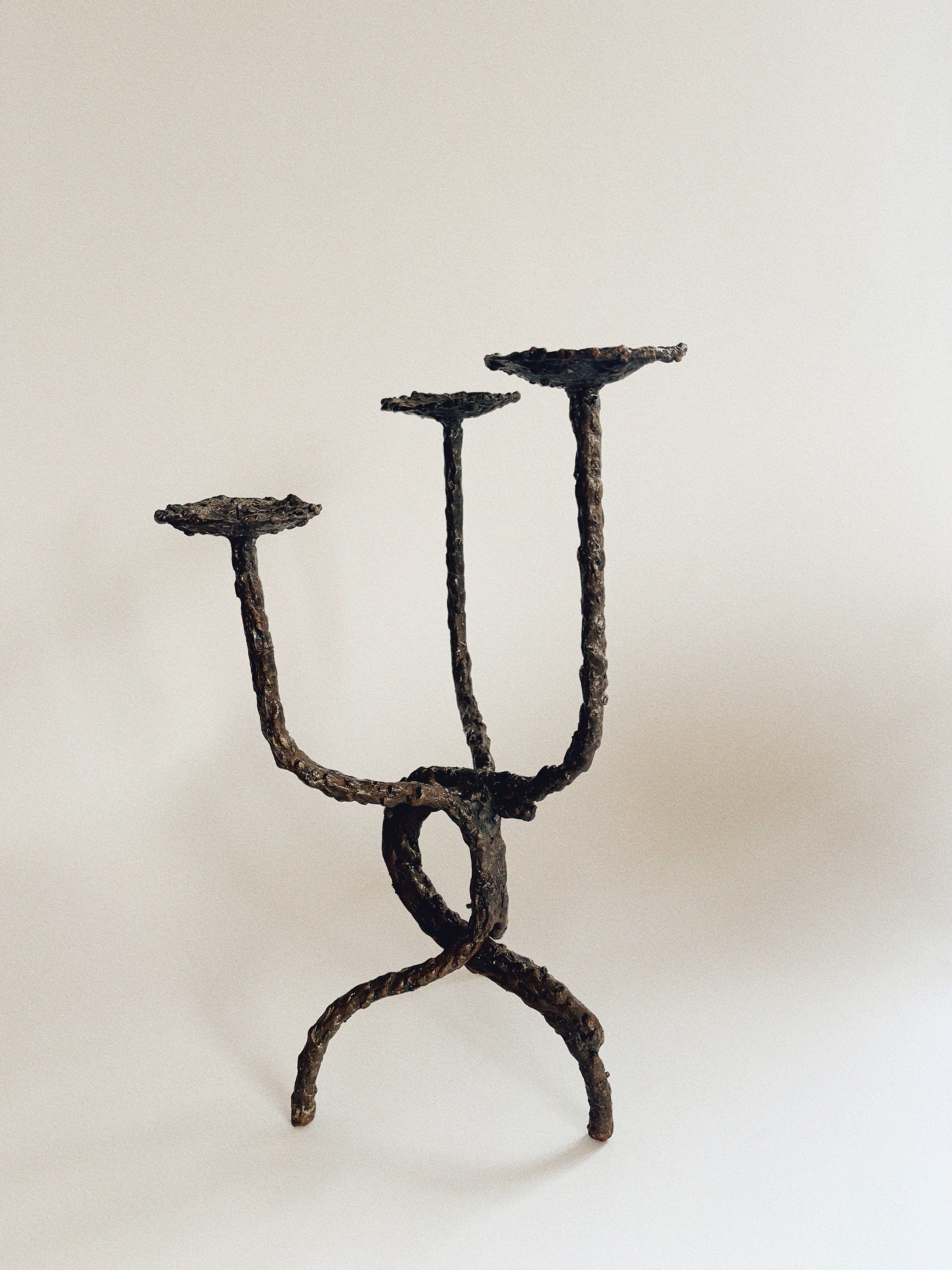 Introducing the Molten Metal Artisan Candlestick by Huerto: a sculptural stand with three branches and a twisted base, reminiscent of a handmade candlestick. Each branch is topped with a flat, circular platform. Its rough texture and appearance of dark, rusted metal create a striking contrast against a plain, light background.