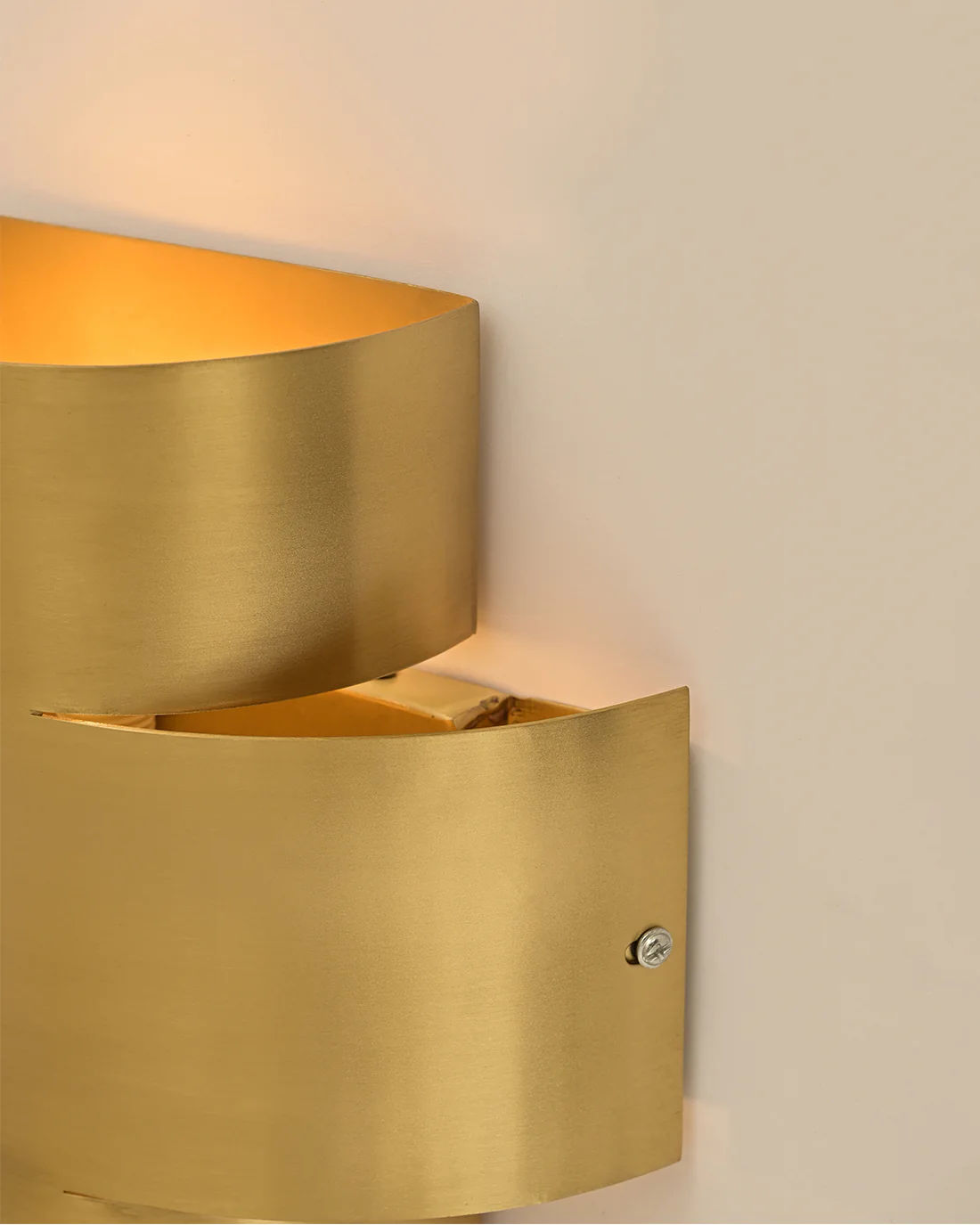 The LMNOH Aura Wall Sconce features a sleek, modern brass design with a minimalist curved shape and visible screw. Emitting soft, warm light, it's ideal for contemporary interiors and creates an elegant contrast against plain beige walls.