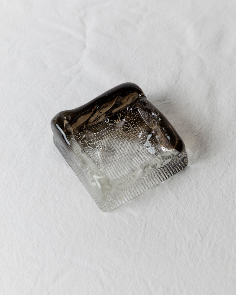 A Vintage Murano Glass Ashtray by Bottega Jacobs, hailing from 1980s Italy, rests on a white textured surface. This small, square glass piece with a dark, smoky edge embodies the Hollywood Regency style with its elegantly textured base and smooth sides, reminiscent of classic Murano glass craftsmanship.