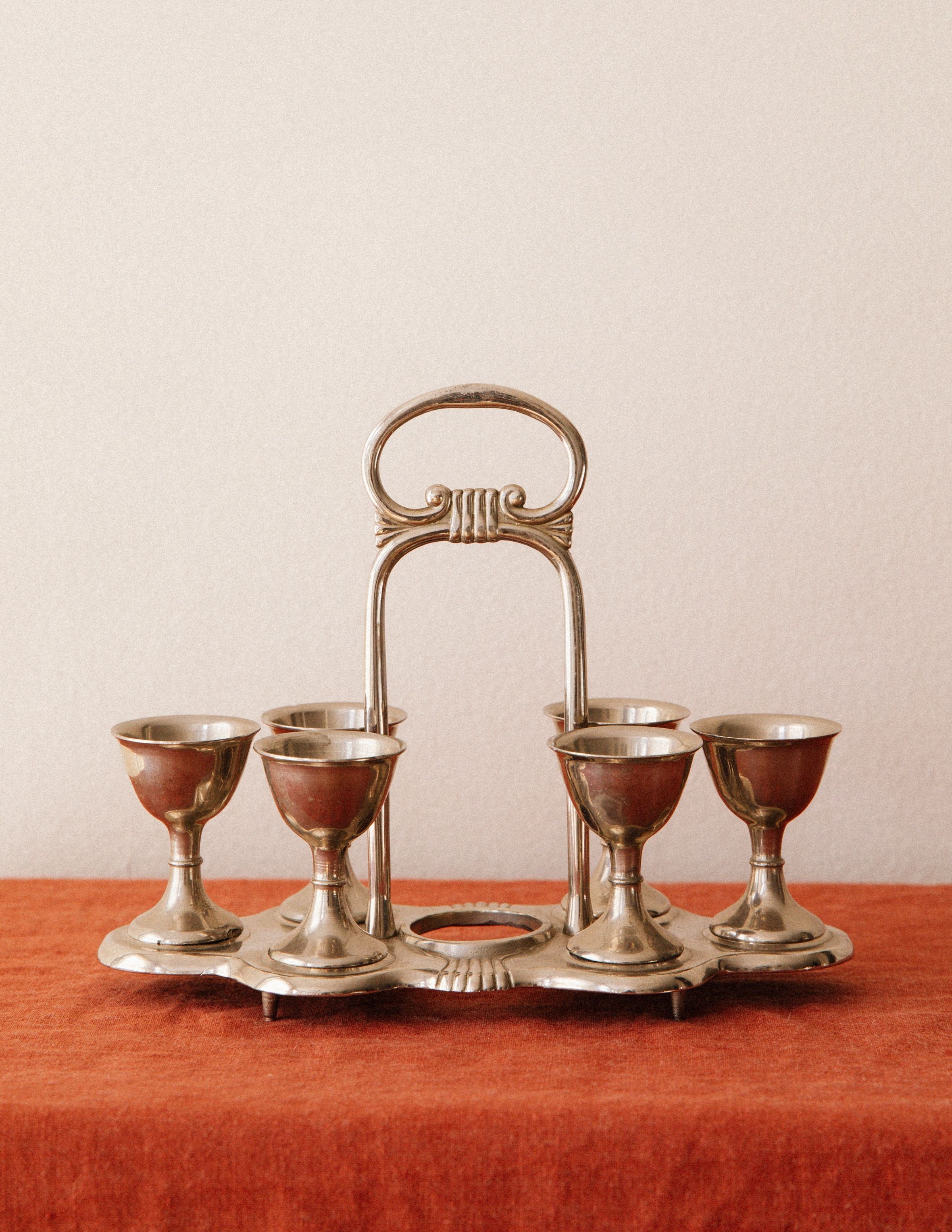 The Arcā Collections Vintage Egg Cruet Holder Set features six sculpted silver-plated cups in a circular display, elegantly presented on a rust-colored cloth before a plain beige wall.
