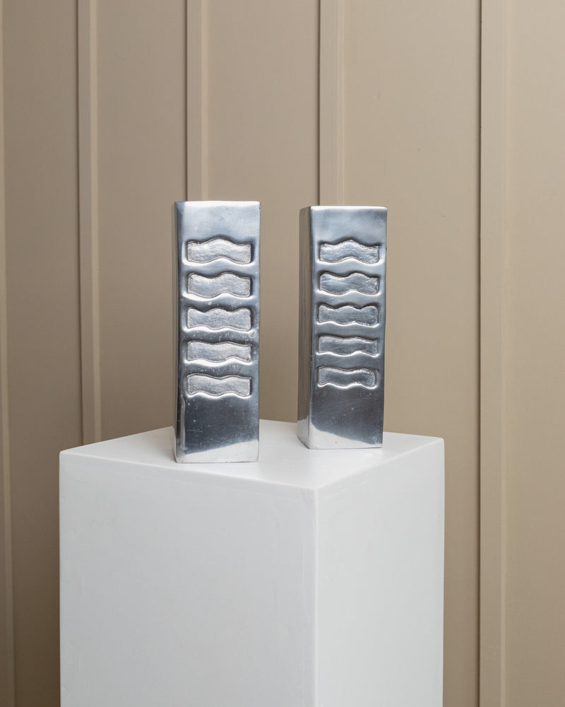 Two geometric metal vases from Bottega Jacobs' Italy 1960s collection, featuring wavy indents, are displayed on a white pedestal. Against a light brown paneled wall, they create a striking visual contrast.