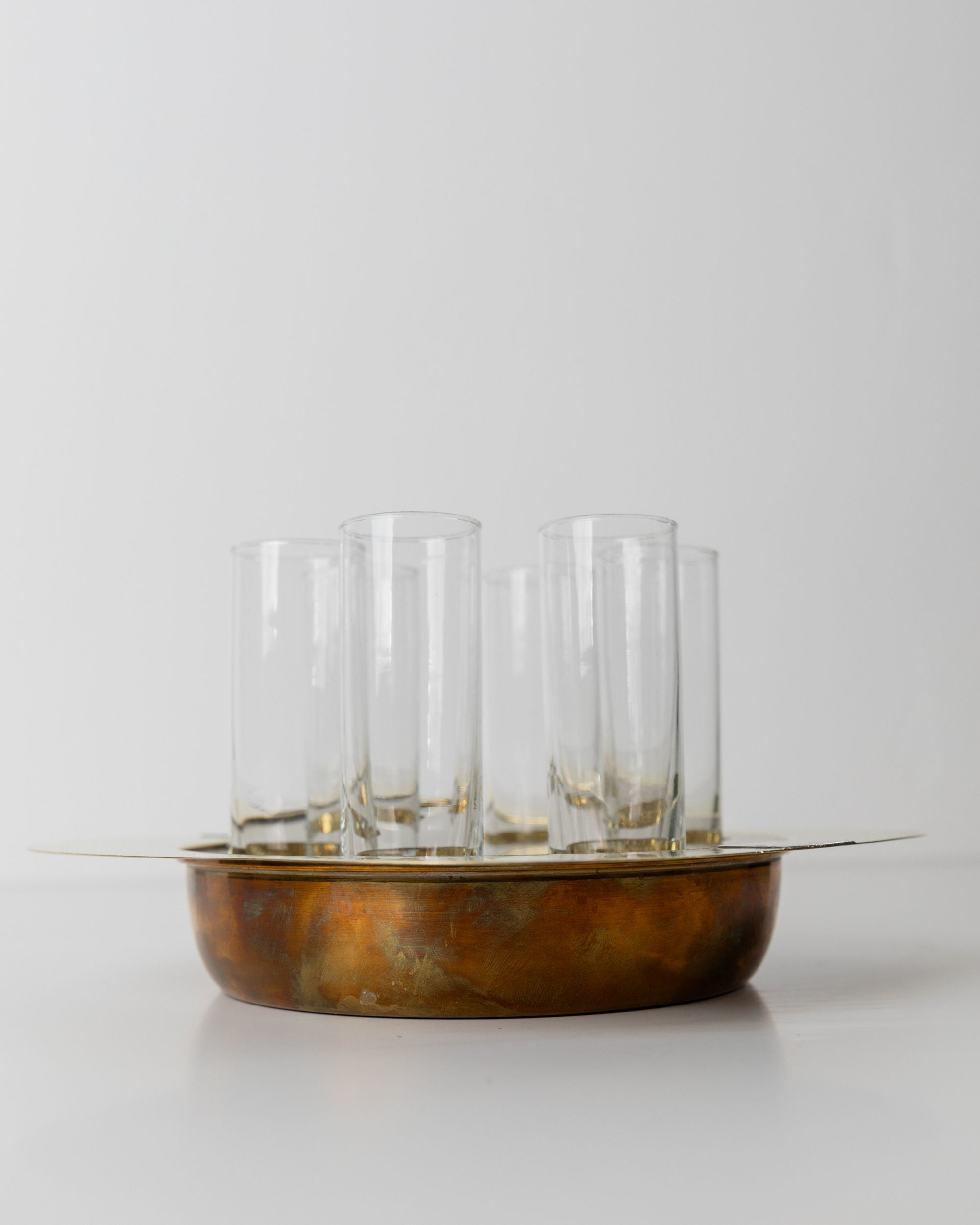 On a white surface, the Set Shot Glasses and Tray - Mid Century Design 70s by Bottega Jacobs holds vintage shot glasses. The slightly worn, round rustic brass tray adds charm, while the uniformly sized glasses are neatly clustered, serving as functional items and decorative accents.