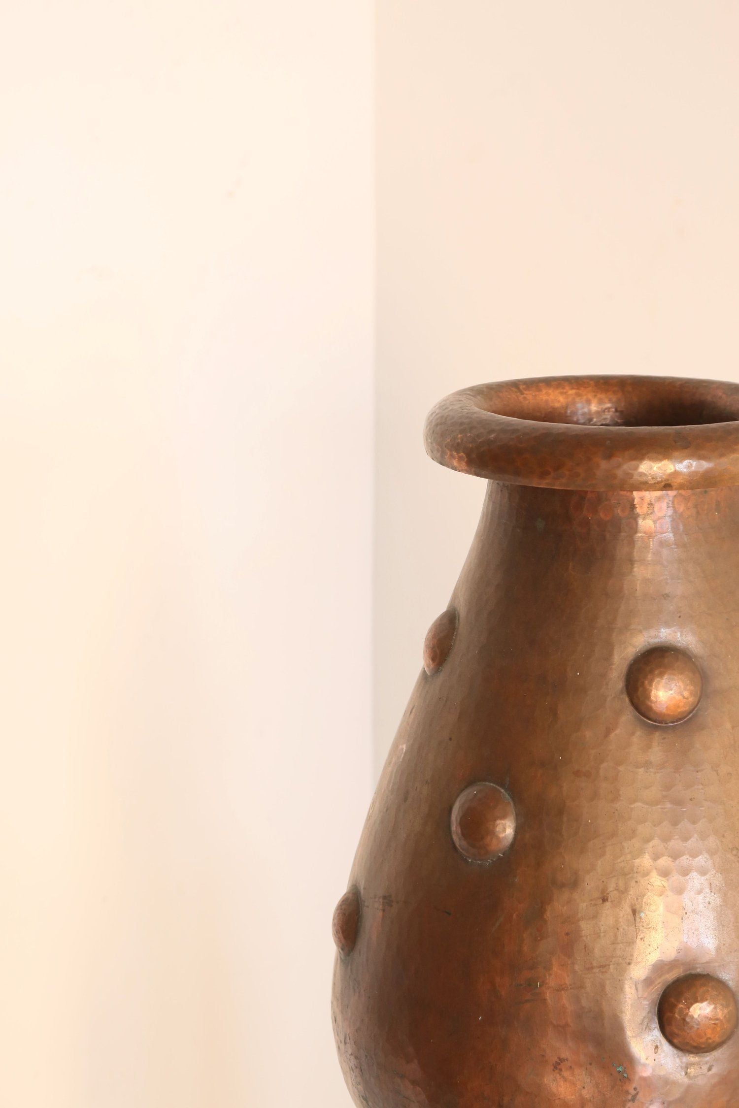 The Hand-Beaten Copper Vase by Ancien et Jolie, featuring circular protrusions, is set against an off-white backdrop near a room's corner. Its wide, slightly flared opening exudes Arts and Crafts charm.