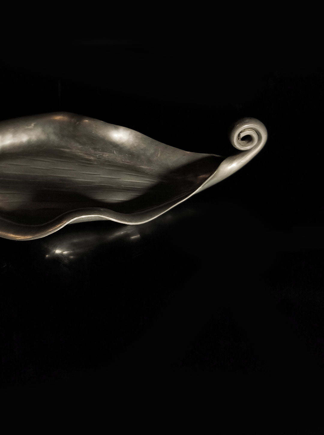 A close-up of the Handmade Leaf Vide Poche by Les Objoies showcases its smooth, wave-like pewter design with a curled tip. This Art Nouveau piece stands against a dark background, accentuating its reflective surface and elegant curves.