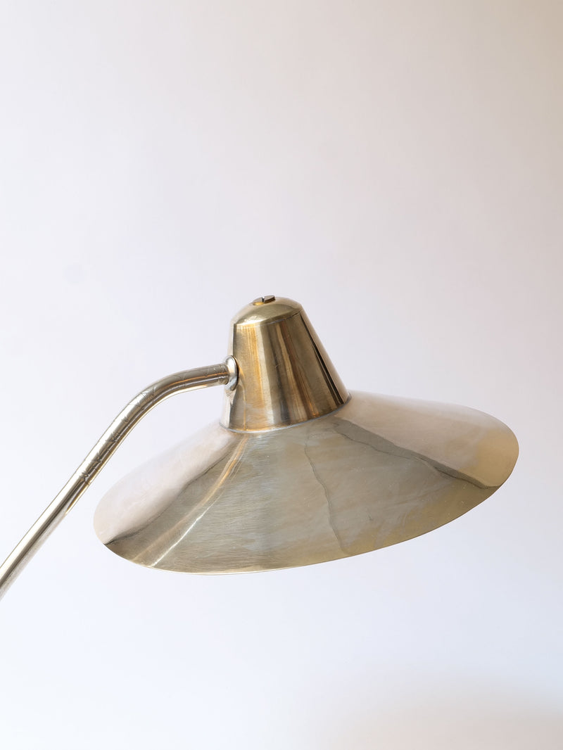 A Silver Brass Desk Lamp from Collection apart with a silver plated brass conical lampshade on a curved stand, set against a plain white background. The lampshade reflects light, highlighting its metallic sheen. The design is minimalist and elegant, showcasing the lamp's sleek and polished appearance.