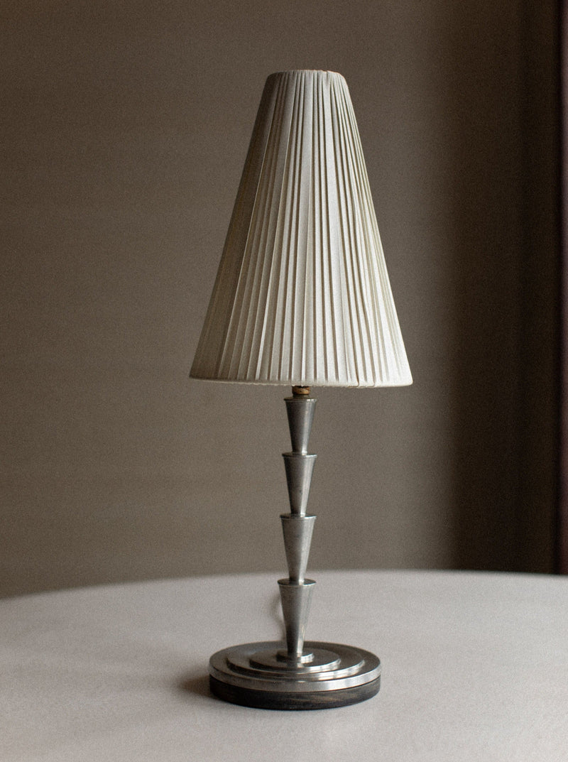 A silver table lamp with a pleated white lampshade stands on a round, metallic base. The modern design, reminiscent of vintage decor, features stacked trapezoidal sections forming the stem. Positioned on a smooth, light-colored surface with a blurred background, this piece echoes the elegance of an Out For Lunch 1920's Swedish Grace Lamp.