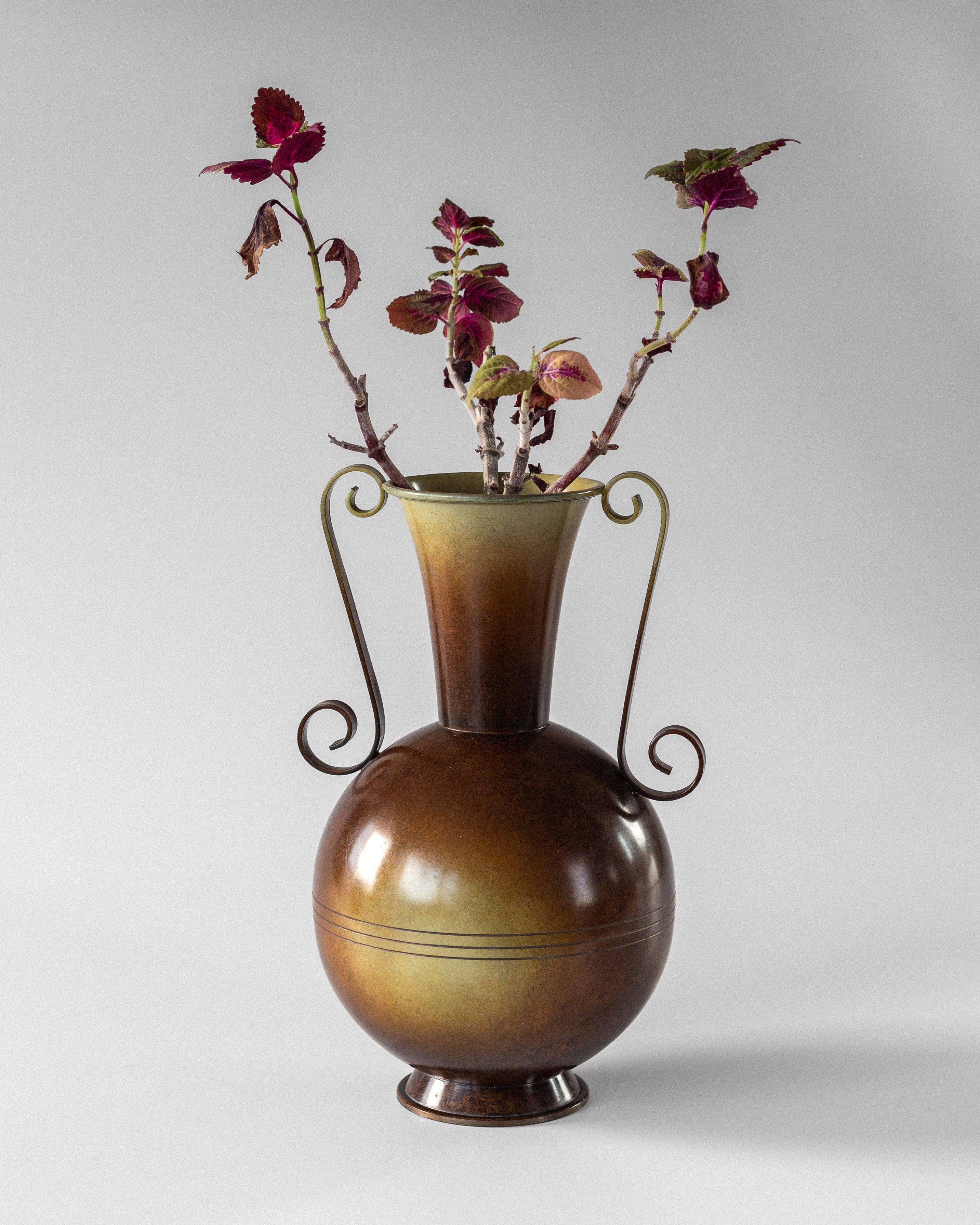 The 1930s Spigel Swedish Art Deco Bronze Vase, with its rounded base, long neck, and elegant curved handles, exudes charm. It holds burgundy leaf branches against a soft gray background that highlights the vase's gradient brown color.