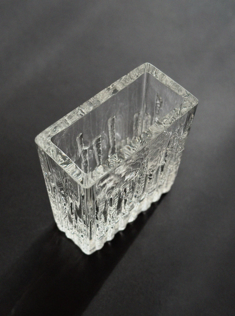 The Vintage Stories Kosta Sweden Ice Glass Vase, featuring vertical ridges and a sculpted, ice-like clear glass surface inspired by Sea Glasbruk designs, rests on a dark matte surface for a minimalist look.
