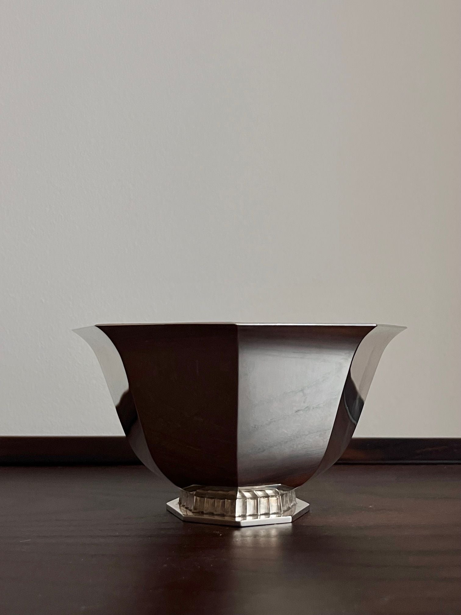 The Art Deco Silver Bowl 1949 by Médecine, featuring a modernist design with sleek angular lines and a hexagonal base, sits on a dark wooden surface. Its reflective finish gives it a minimalist elegance against the plain, off-white background.