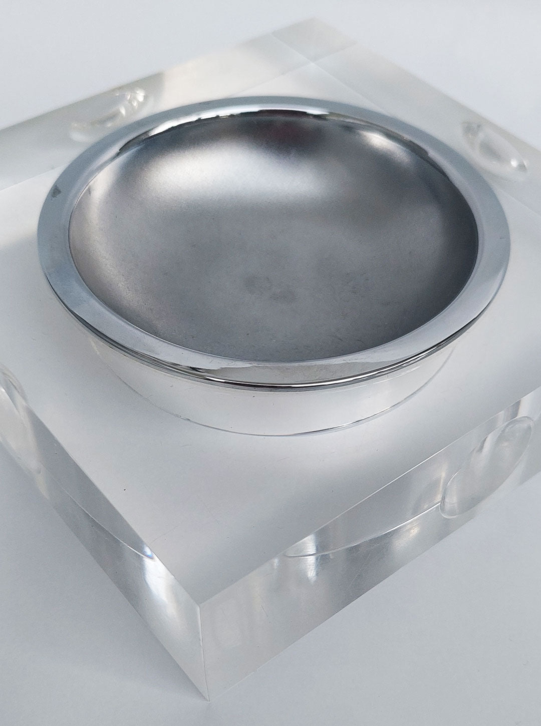 A sleek, modern 70's Plexiglass Ashtray by Les Objoies with a polished metallic finish is set on a clear, cubic base. Drawing inspiration from space-age aesthetics, the clean, minimalist design emphasizes the reflective surface of the ashtray and the transparent block it rests on. The background is a neutral light gray.