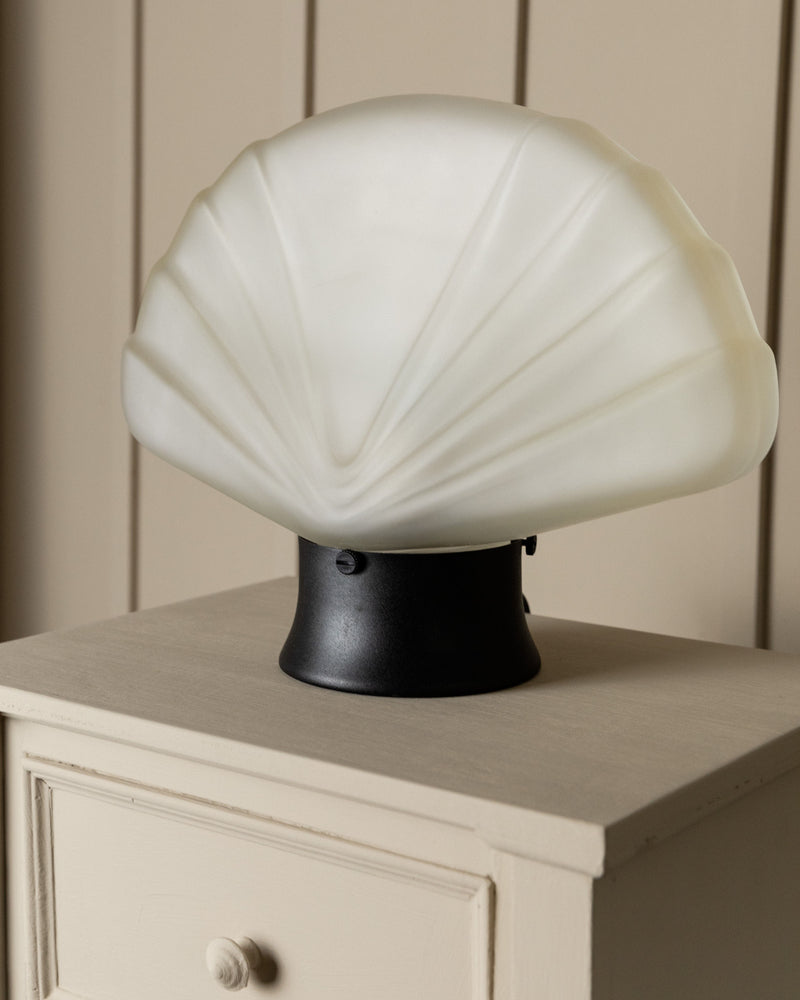 A Murano Glass Table Lamp from the 80s by Bottega Jacobs, featuring a vintage design with a shell-shaped white top and black base, sits on a beige wooden nightstand with a knobbed drawer, nestled against a beige paneled wall.