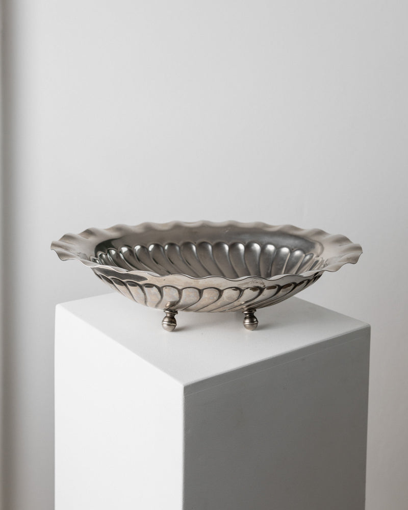 The Bottega Jacobs Large Silver-plated Bowl from Italy, 1940s features a vintage design with a fluted, scalloped edge and sits elegantly on a white pedestal. Its three small ball-shaped feet add an elegant lift, making it a standout centerpiece against the plain white wall backdrop.