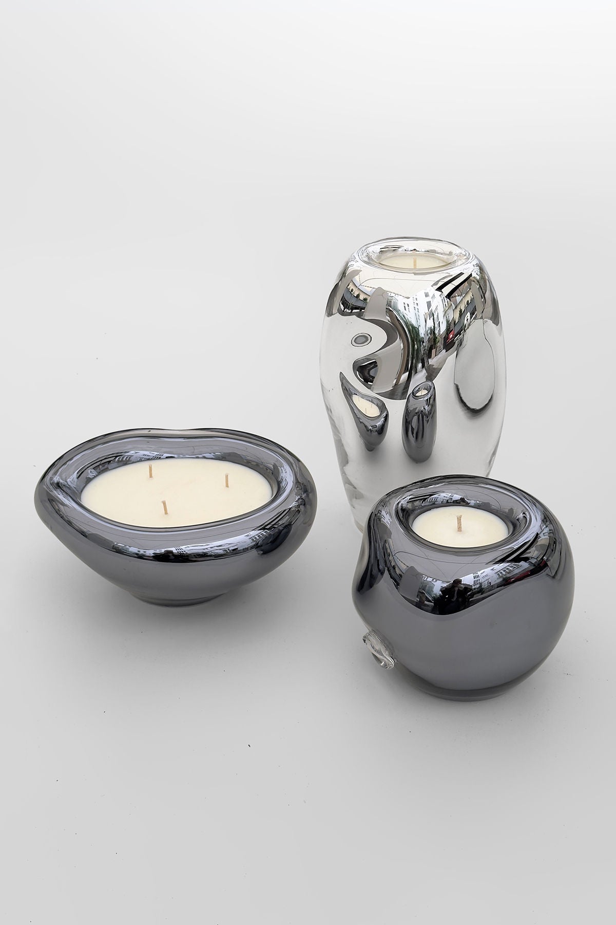 Three Lumi Candles by HOMA from the LUMI Collection are on a white surface: one is tall and shiny like a Silver Mirror with three wicks, another is rounded and metallic gray with one wick, and the last is low, oval-shaped, and metallic gray with two wicks.