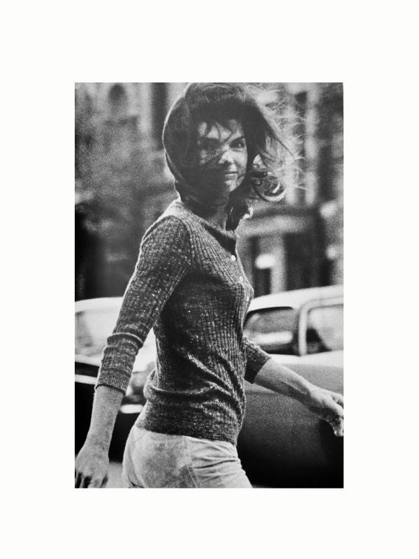 Black and white photograph of a woman walking outdoors. She is wearing a sweater and pants, with her hair blowing in the wind. The background includes parked cars and buildings, capturing a moment reminiscent of *Happy Times by Lee Radziwill (Signed Copy)* from Maison Plage in personal archives.