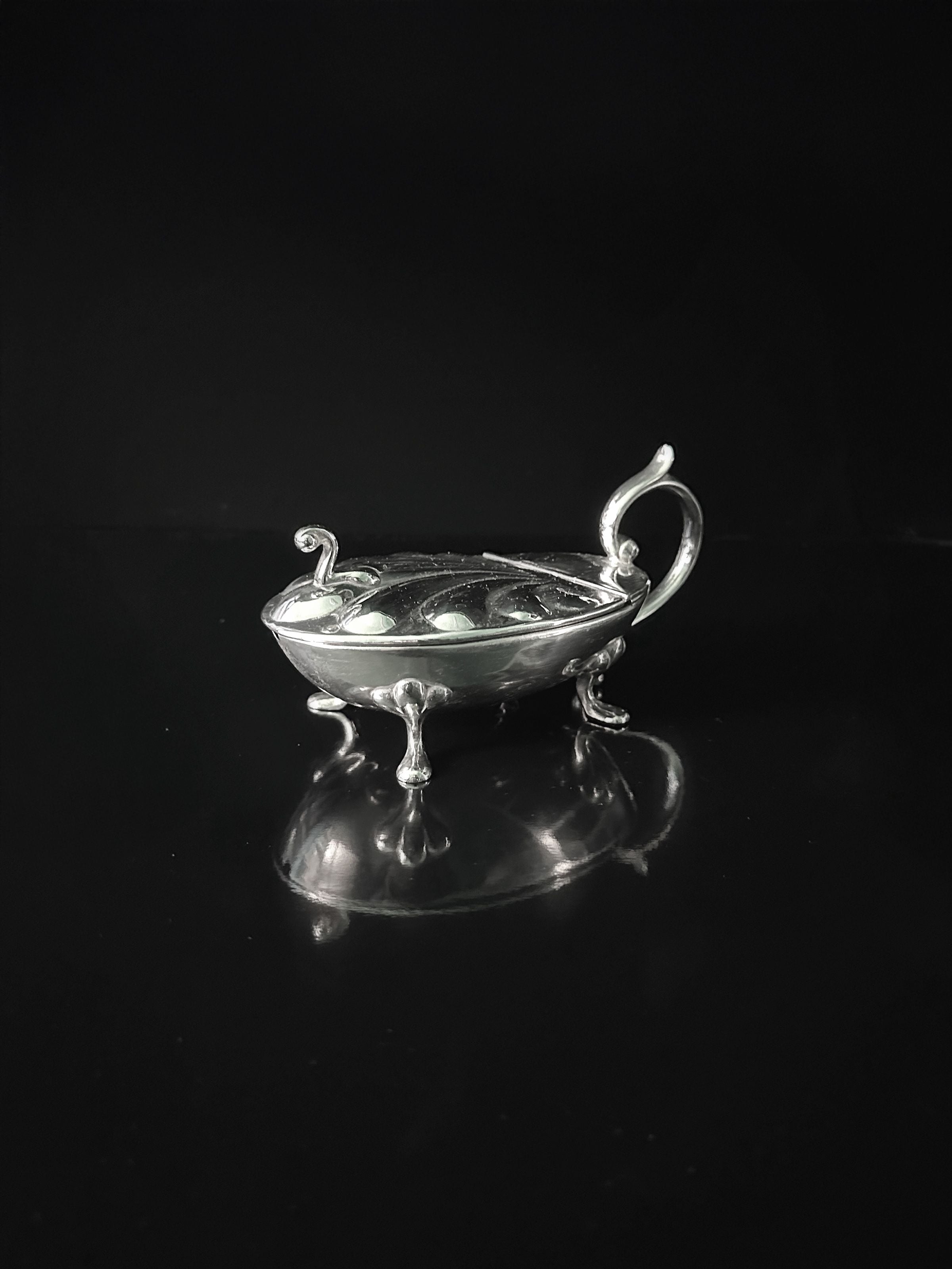 The Silver-plated Caviar Frog 1950s by Dodo Vintage is a small, ornate container with intricate handles and four decorative legs. It resembles a frog-shaped figure on a reflective black surface, showcasing detailed craftsmanship with its smooth, polished finish.