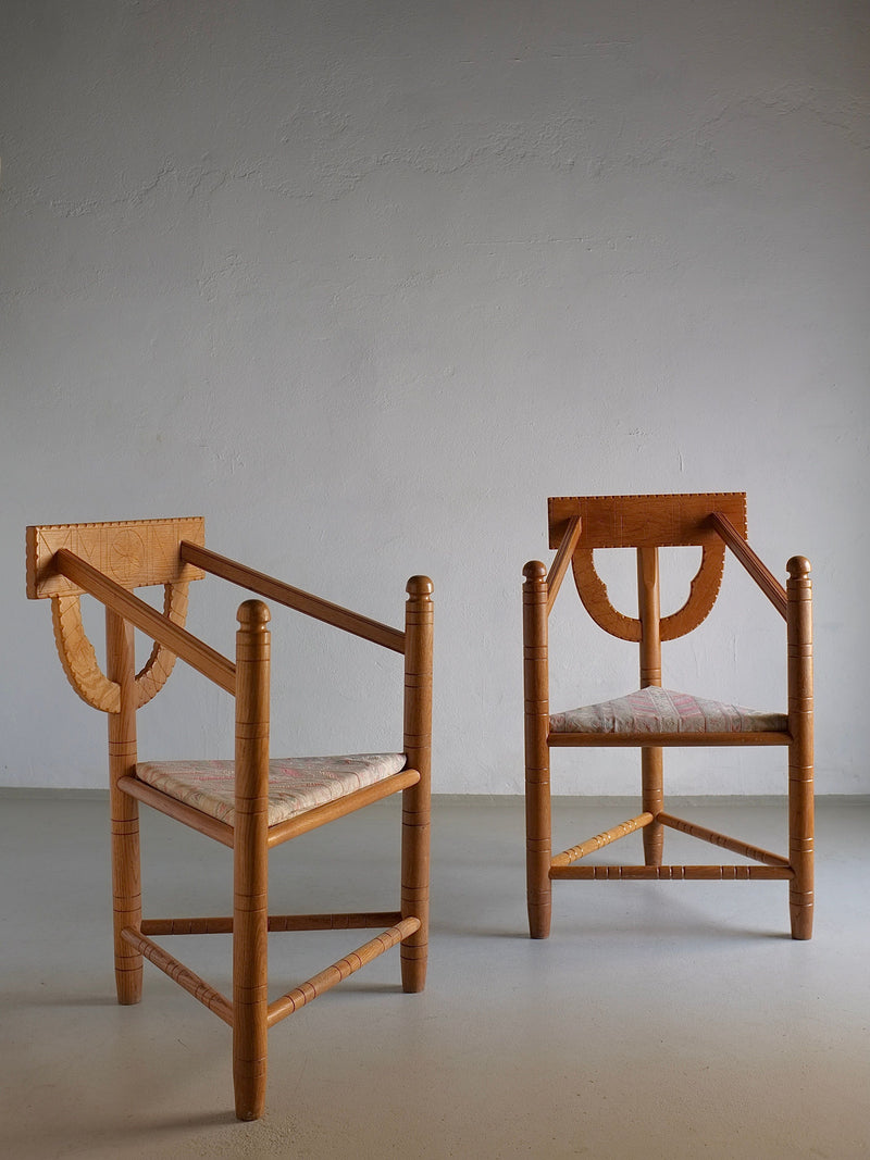 Two Rustic Carved Oak Corner Chairs by Veter Vintage are positioned against a plain, textured wall. Both chairs feature unique triangular and curved designs on their backrests, reminiscent of Sweden's design period in the 1950s. They have light-colored, cushioned seats and are placed on a smooth, light gray floor.